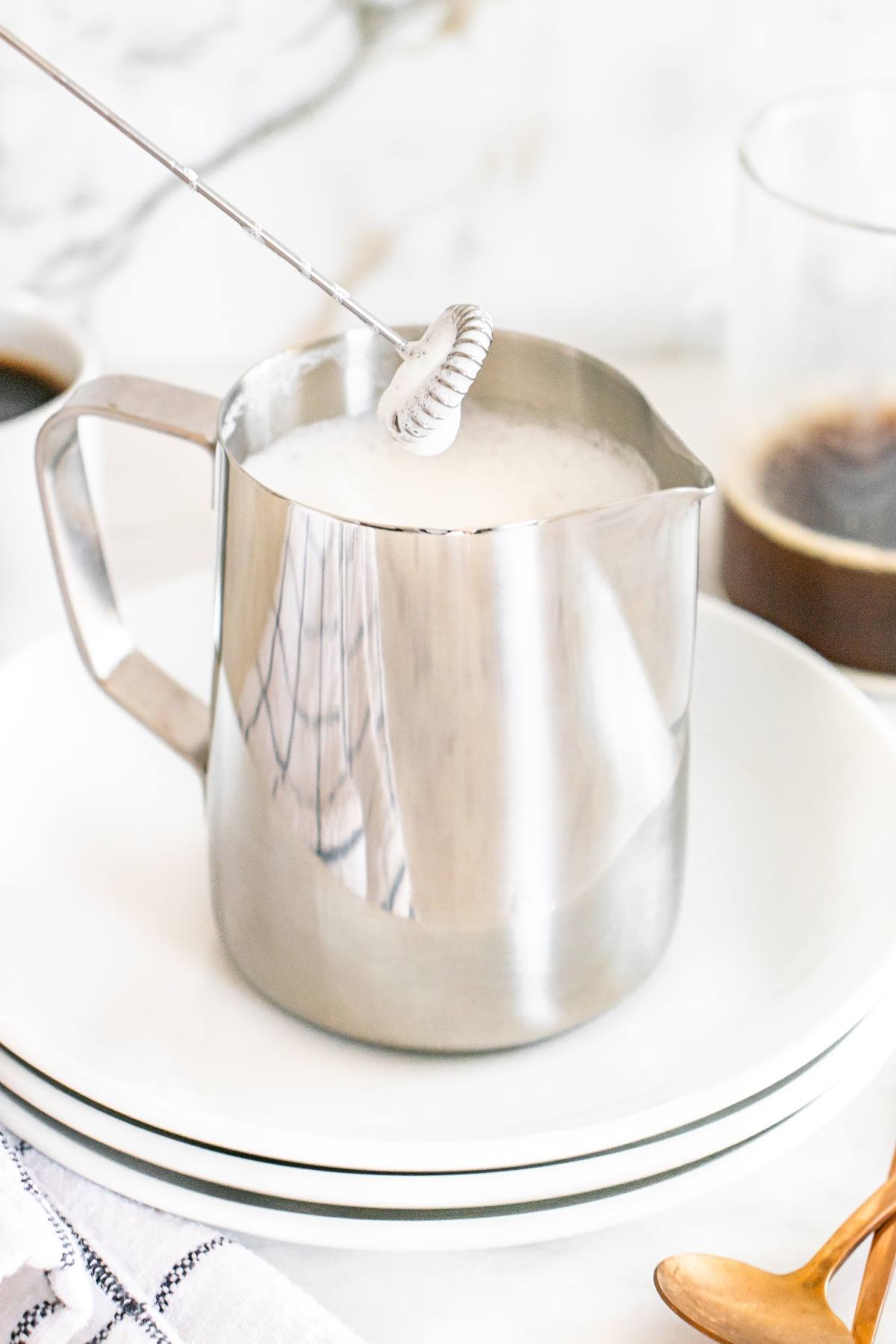 A frother whipping milk in a metal pitcher on a white plate, with coffee and a spoon nearby.