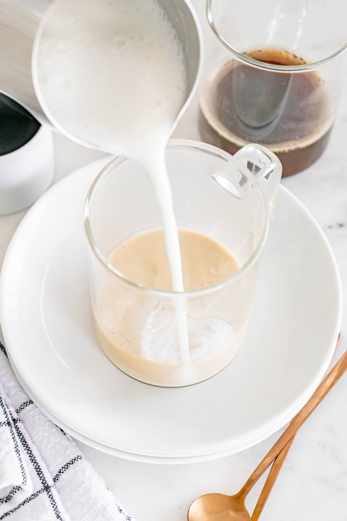 Milk is being poured into a clear glass mug with coffee, on a white plate with a copper spoon nearby.