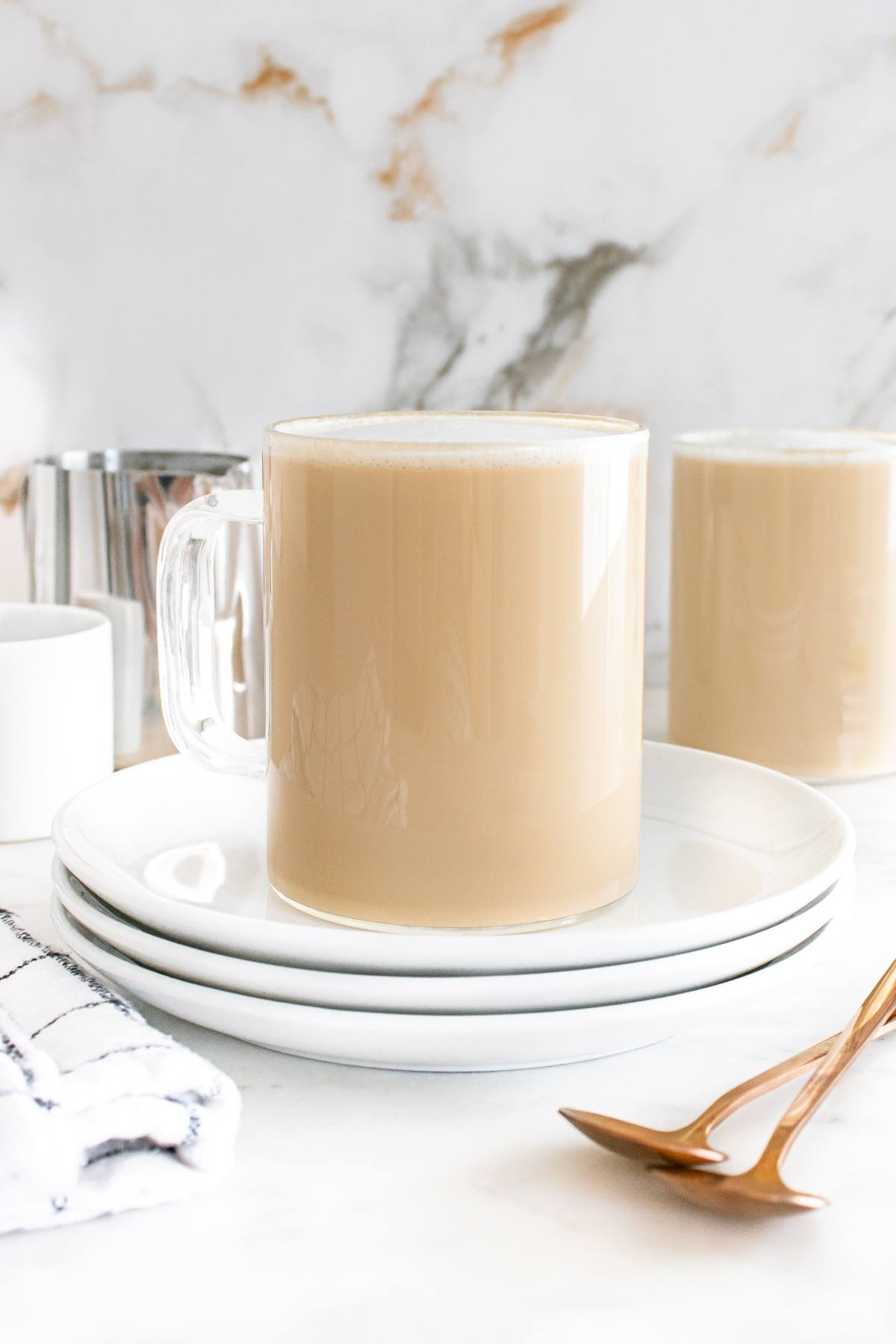A glass mug of coffee with milk on stacked white plates, next to a napkin and gold spoons.