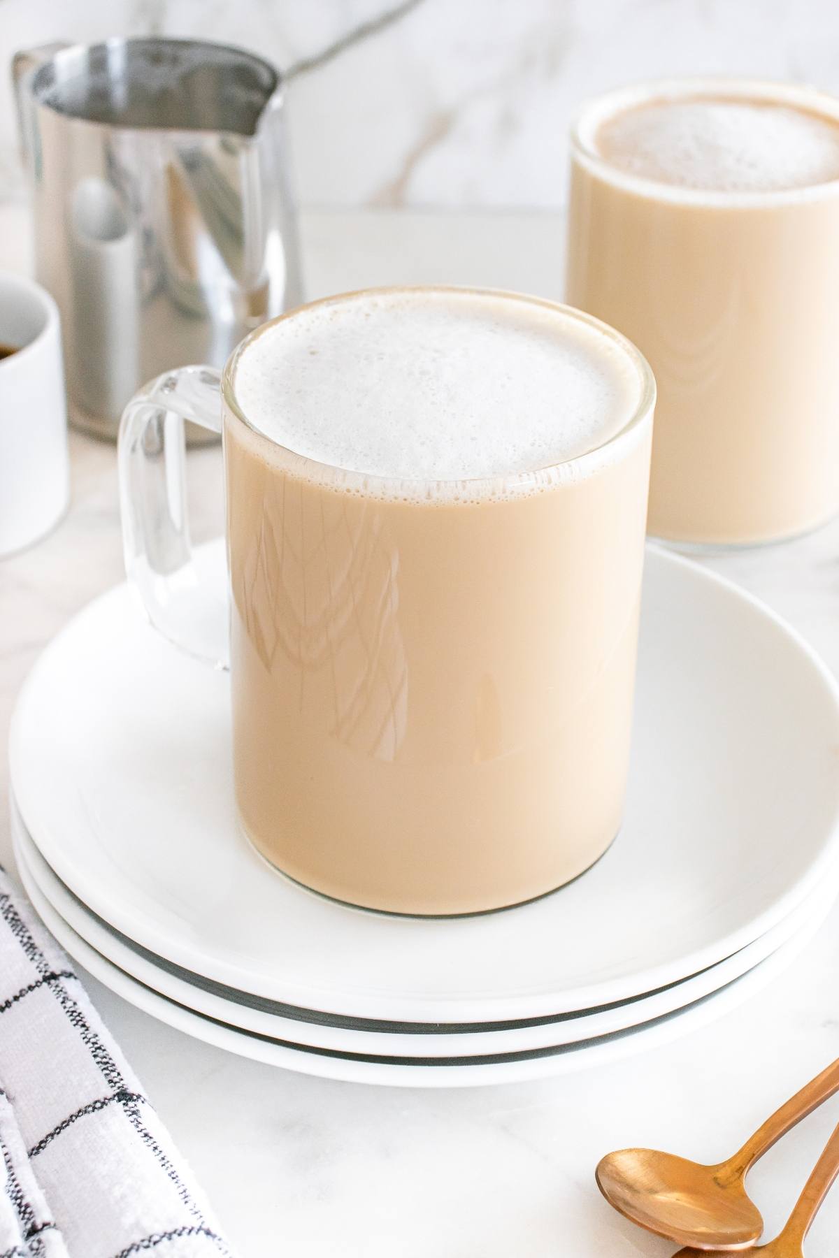 A glass mug of frothy chai latte on a white plate, with another mug and a gold spoon in the background.