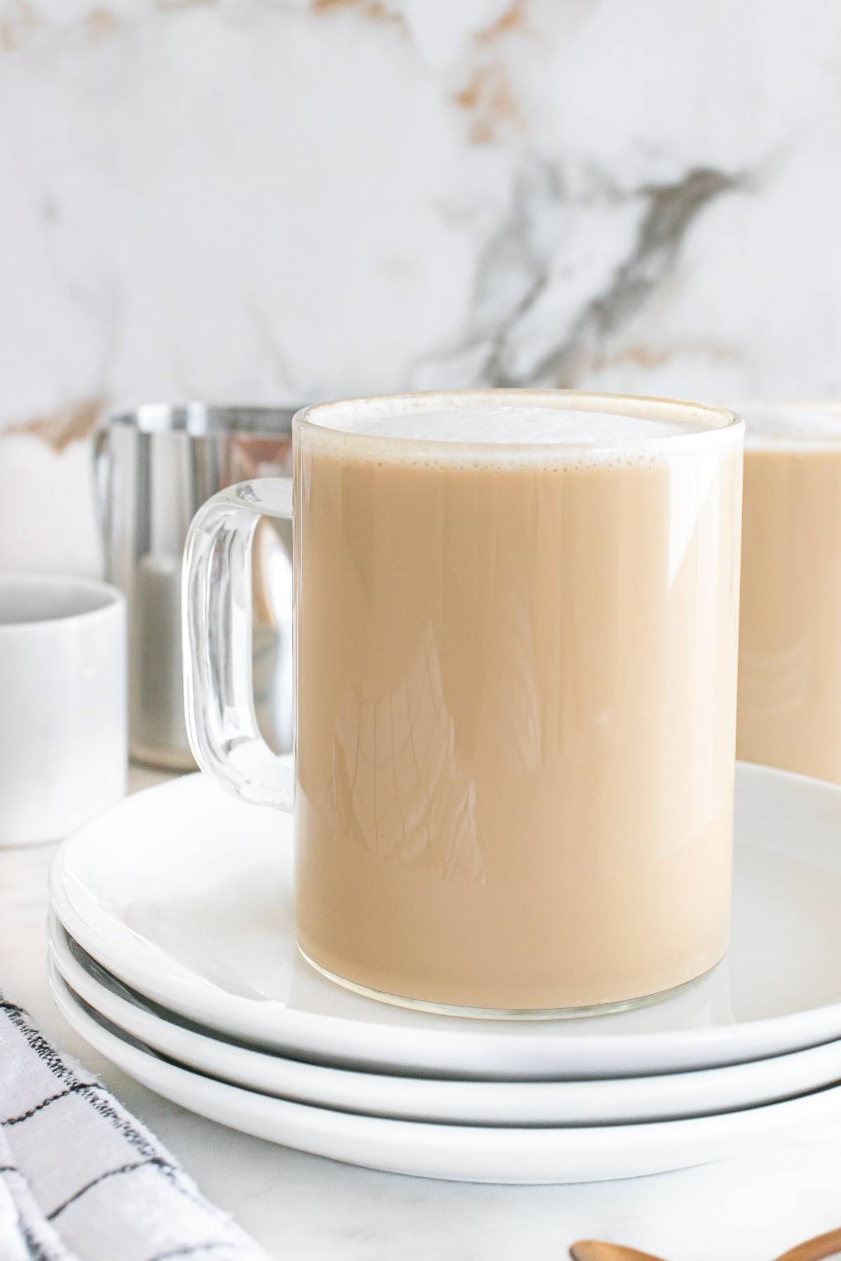 A glass mug of creamy milk tea sits on stacked white plates with a soft, marble background.