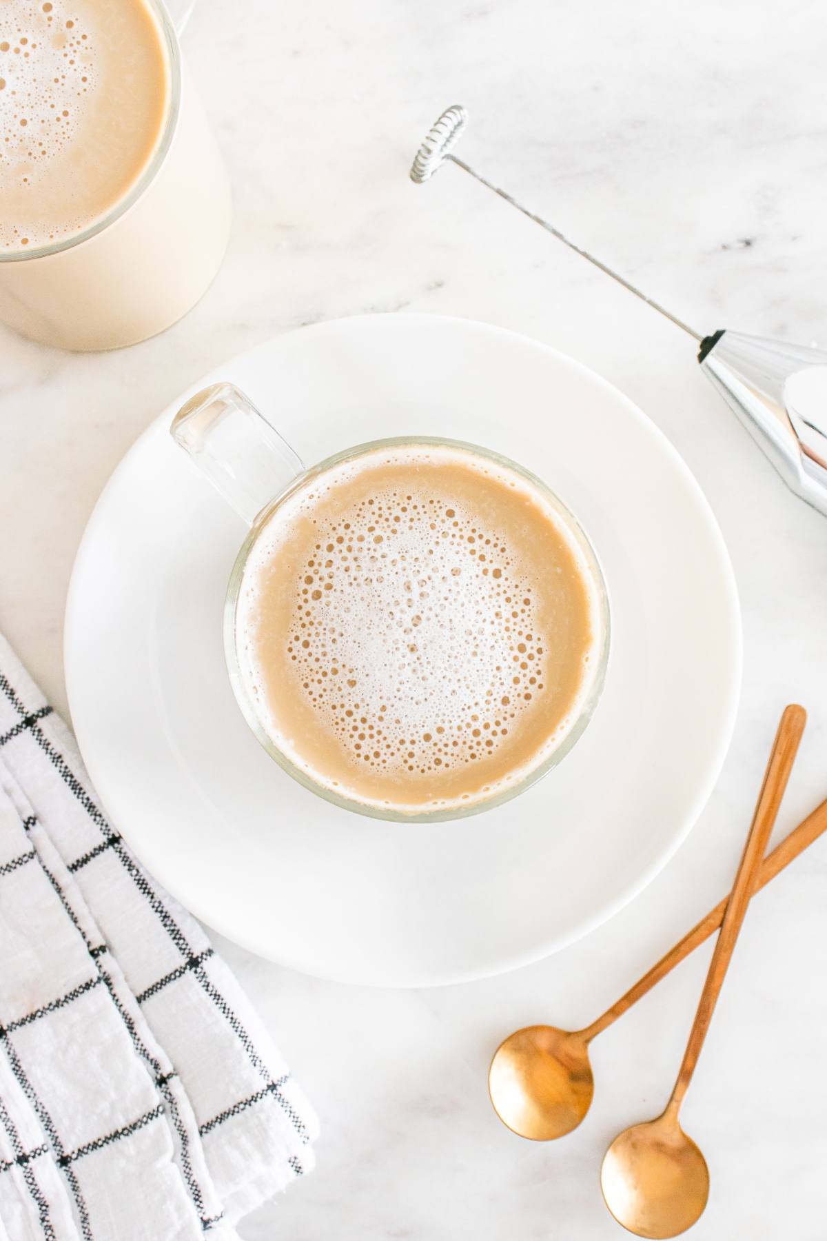A cup of frothy coffee on a white plate with two gold spoons, a towel, and a milk frother on a marble surface.