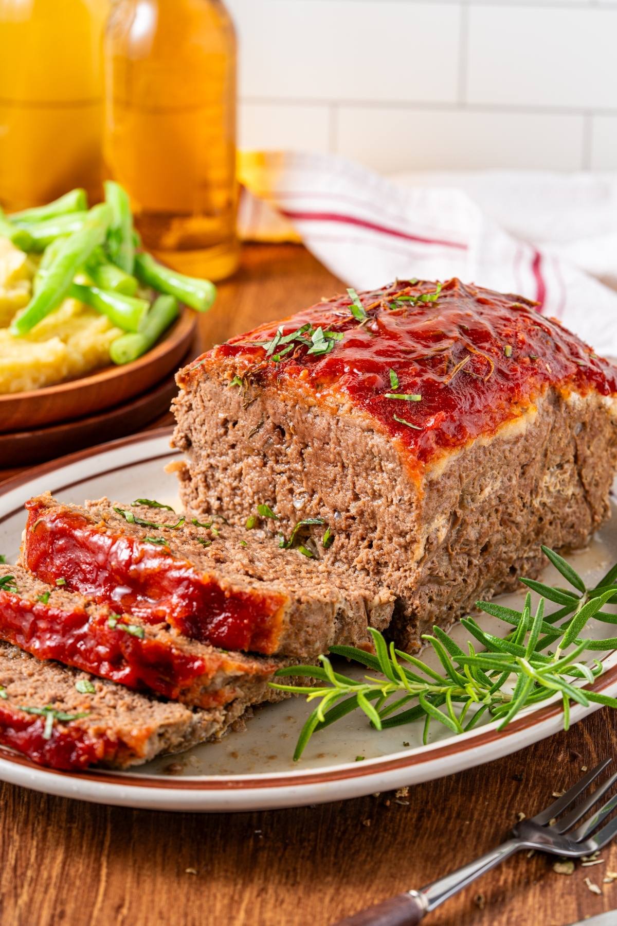 A glazed meatloaf sliced on a plate, garnished with herbs, with green beans and potatoes in the background.