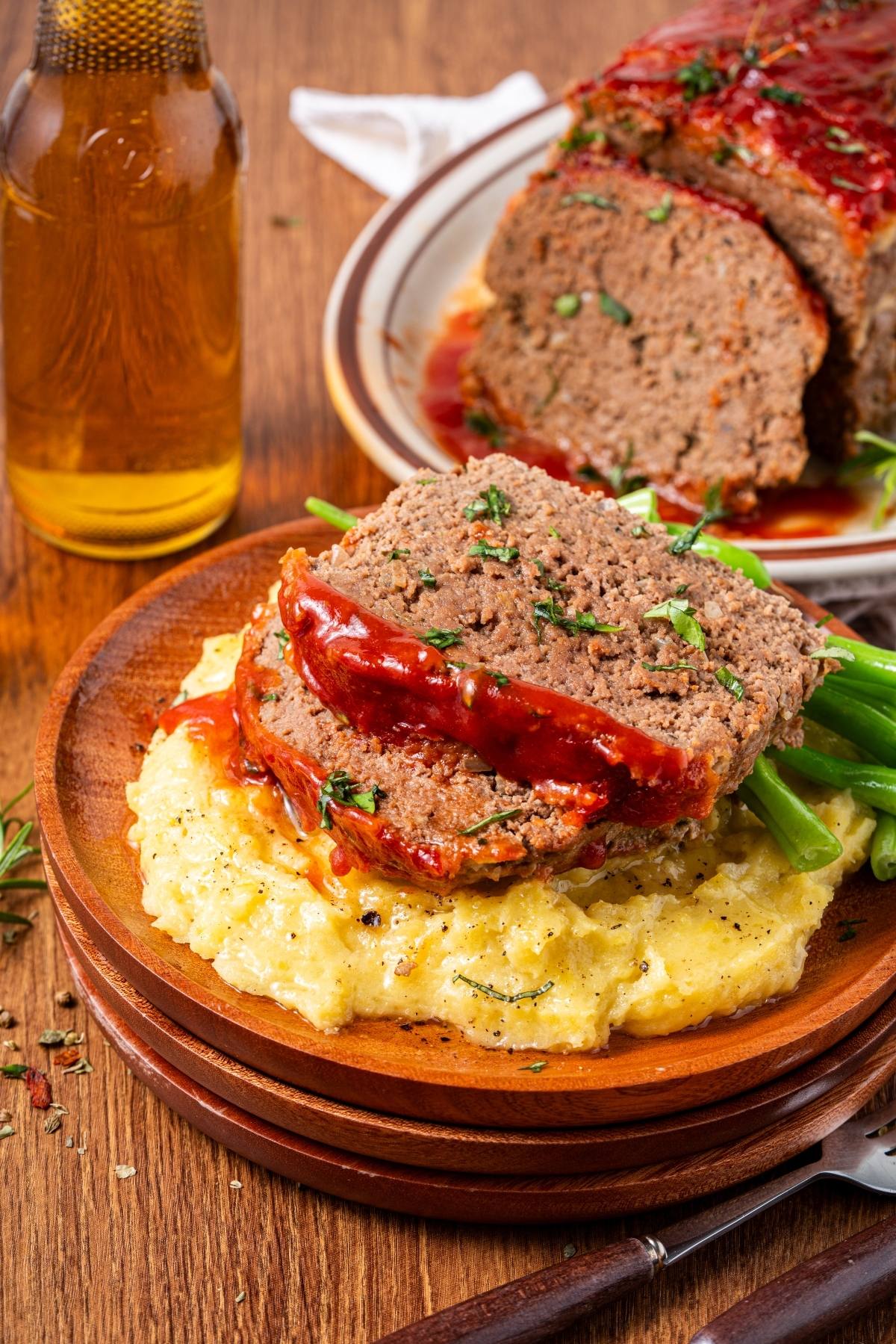 Slices of glazed meatloaf on mashed potatoes with green beans, served on a wooden plate.