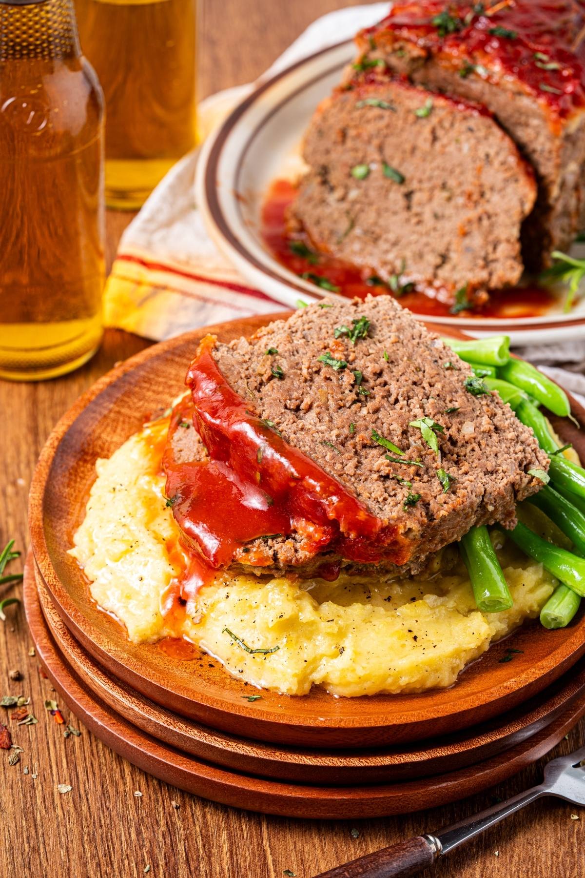 Two slices of meatloaf with ketchup on mashed potatoes and green beans, served on a wooden plate.