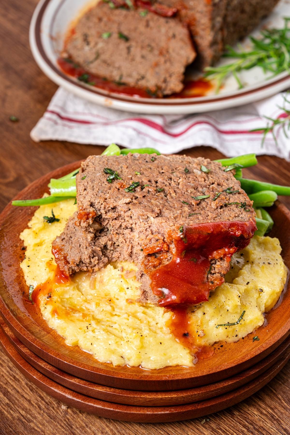 Slice of meatloaf with gravy served on cheesy mashed potatoes and green beans on a wooden plate.
