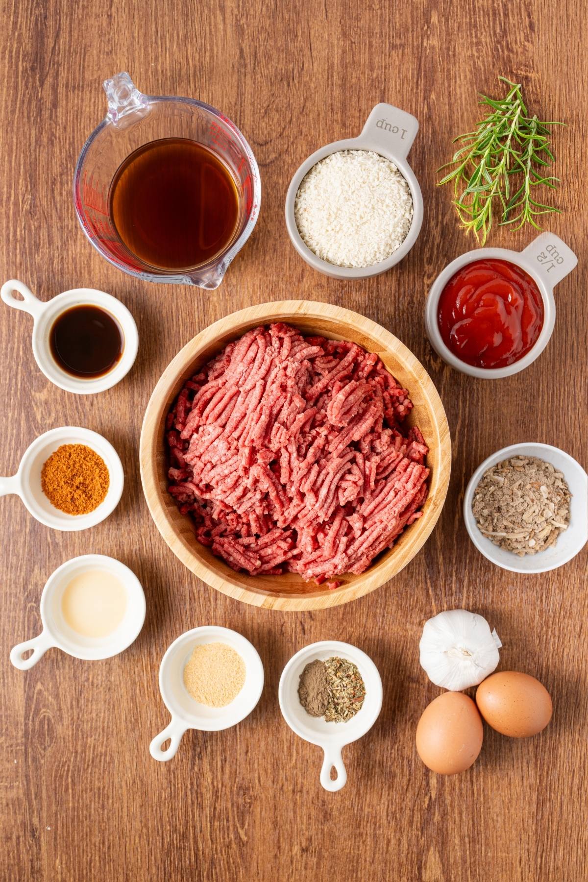 Raw ground beef, eggs, seasonings, sauces, breadcrumbs, and herbs arranged on a wooden table, viewed from above.
