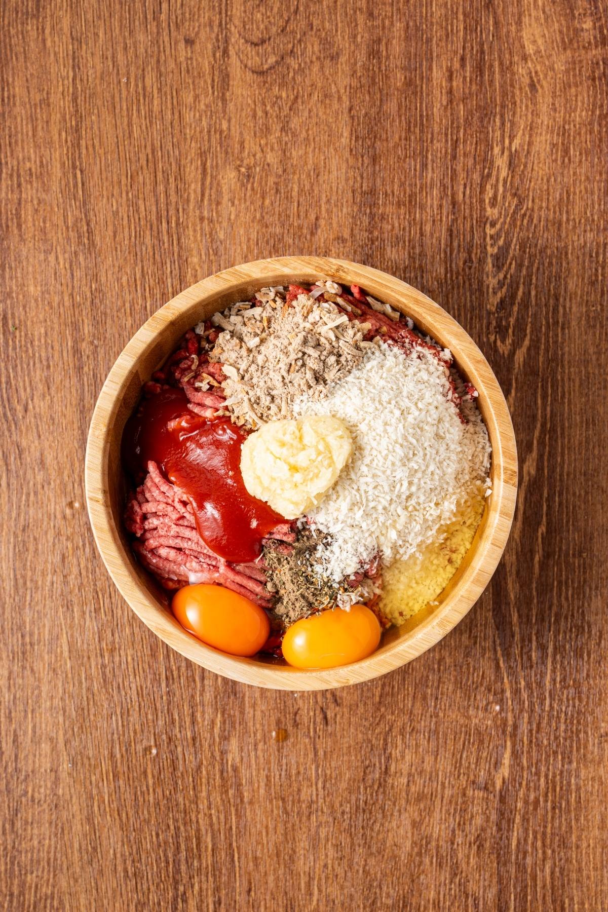 A wooden bowl with ground beef, eggs, ketchup, breadcrumbs, grated onion, and seasonings on a wood surface.