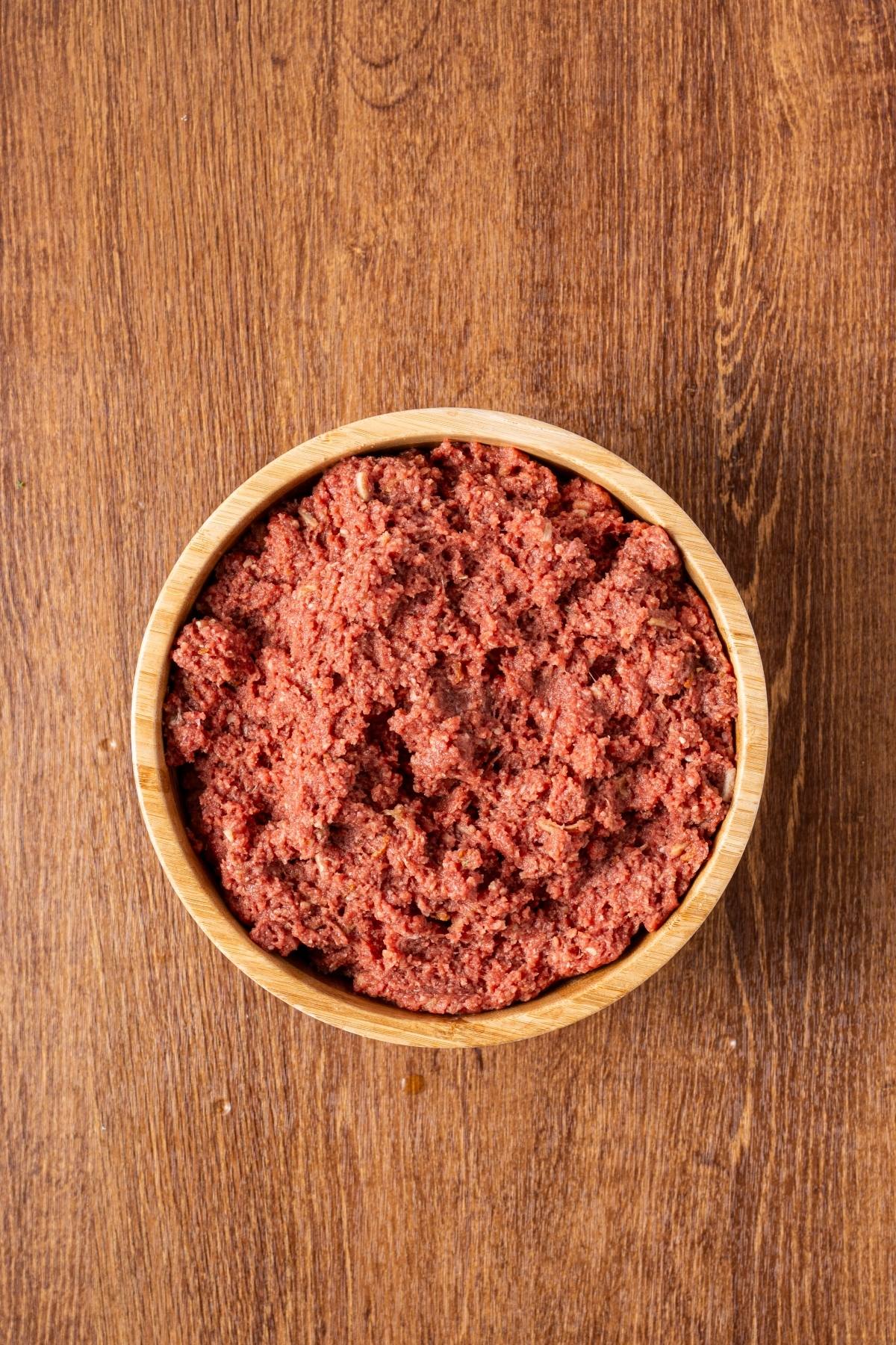 A wooden bowl filled with raw ground beef sits on a wooden surface.