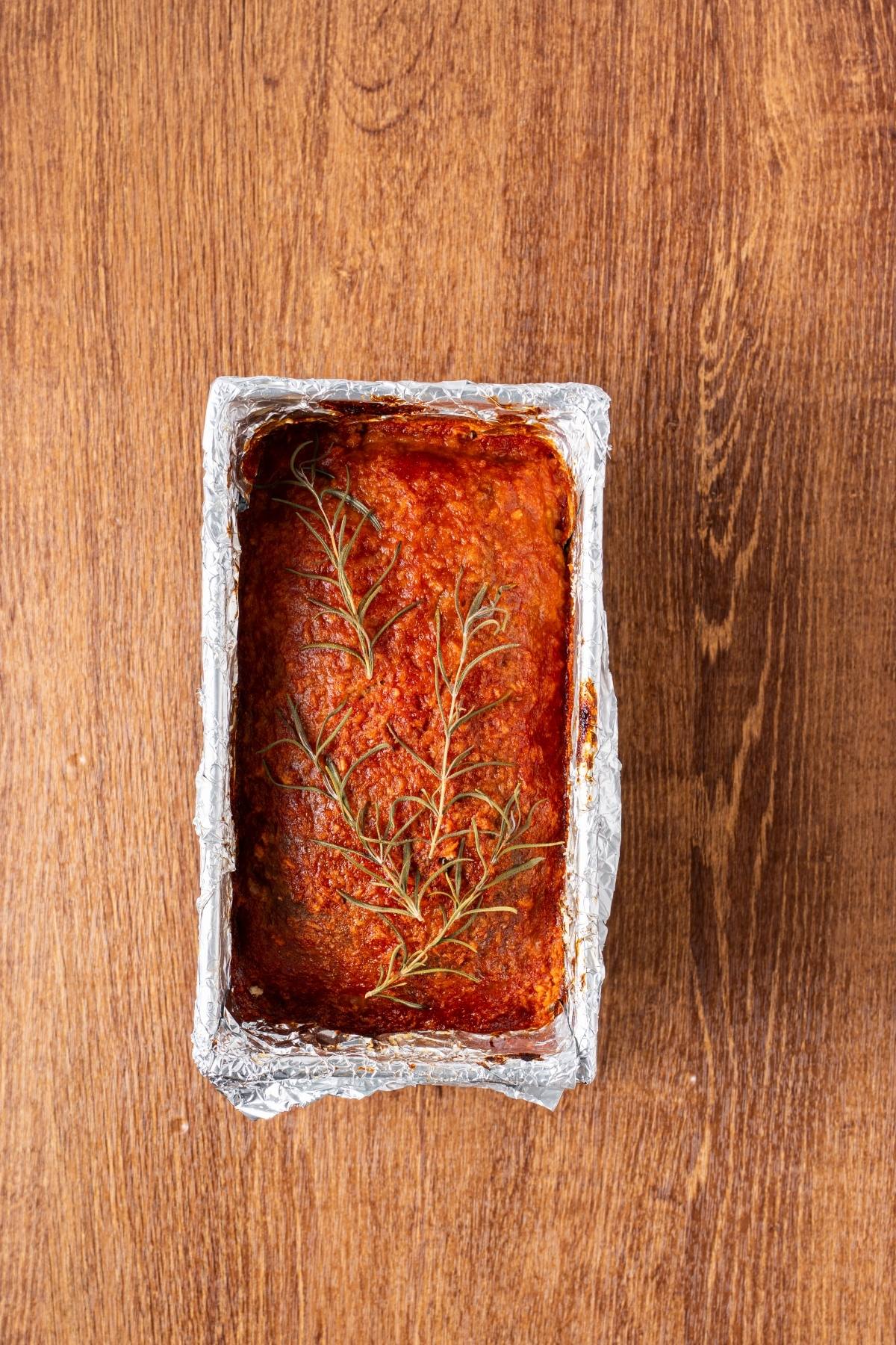 A baked meatloaf in a foil-lined pan, topped with sprigs of rosemary, on a wooden surface.