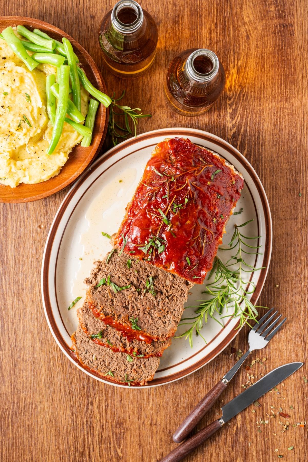 Sliced meatloaf on a plate with herbs, next to mashed potatoes and green beans on a wooden table.