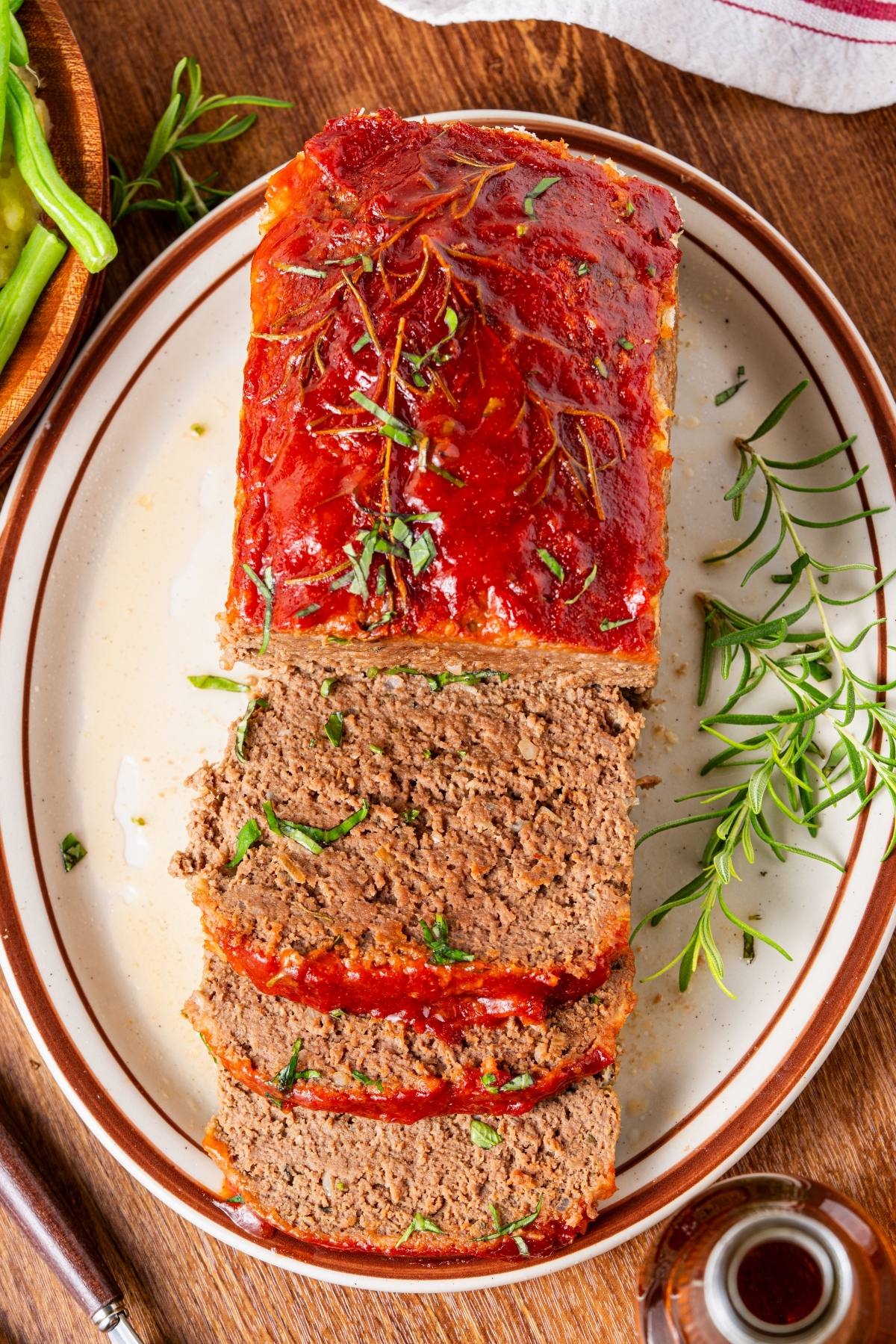 A sliced meatloaf topped with ketchup and herbs on a plate, with fresh rosemary on the side.