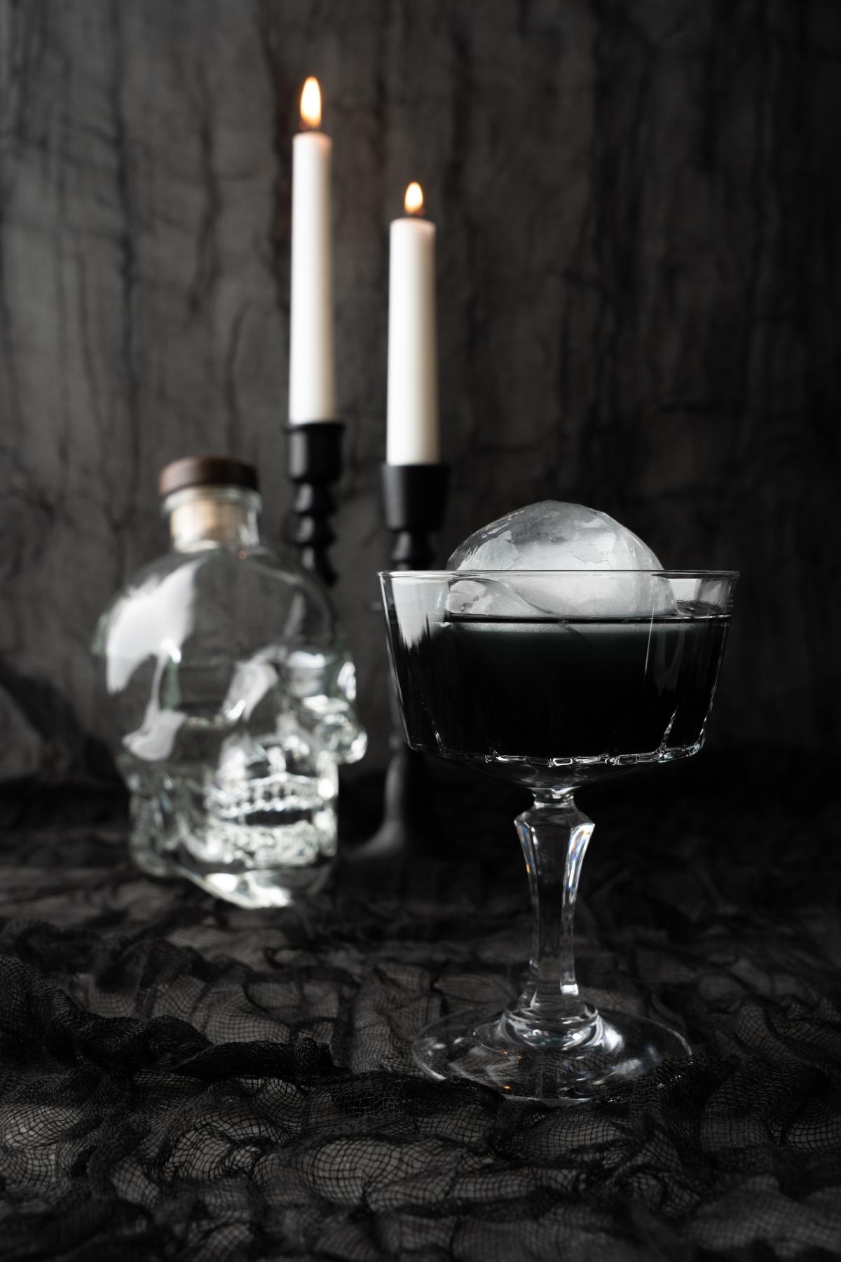A black cocktail served over a round ice cube in a glass sits beside a skull-shaped bottle, with two lit candles glowing in the background—a perfect Halloween themed drink.