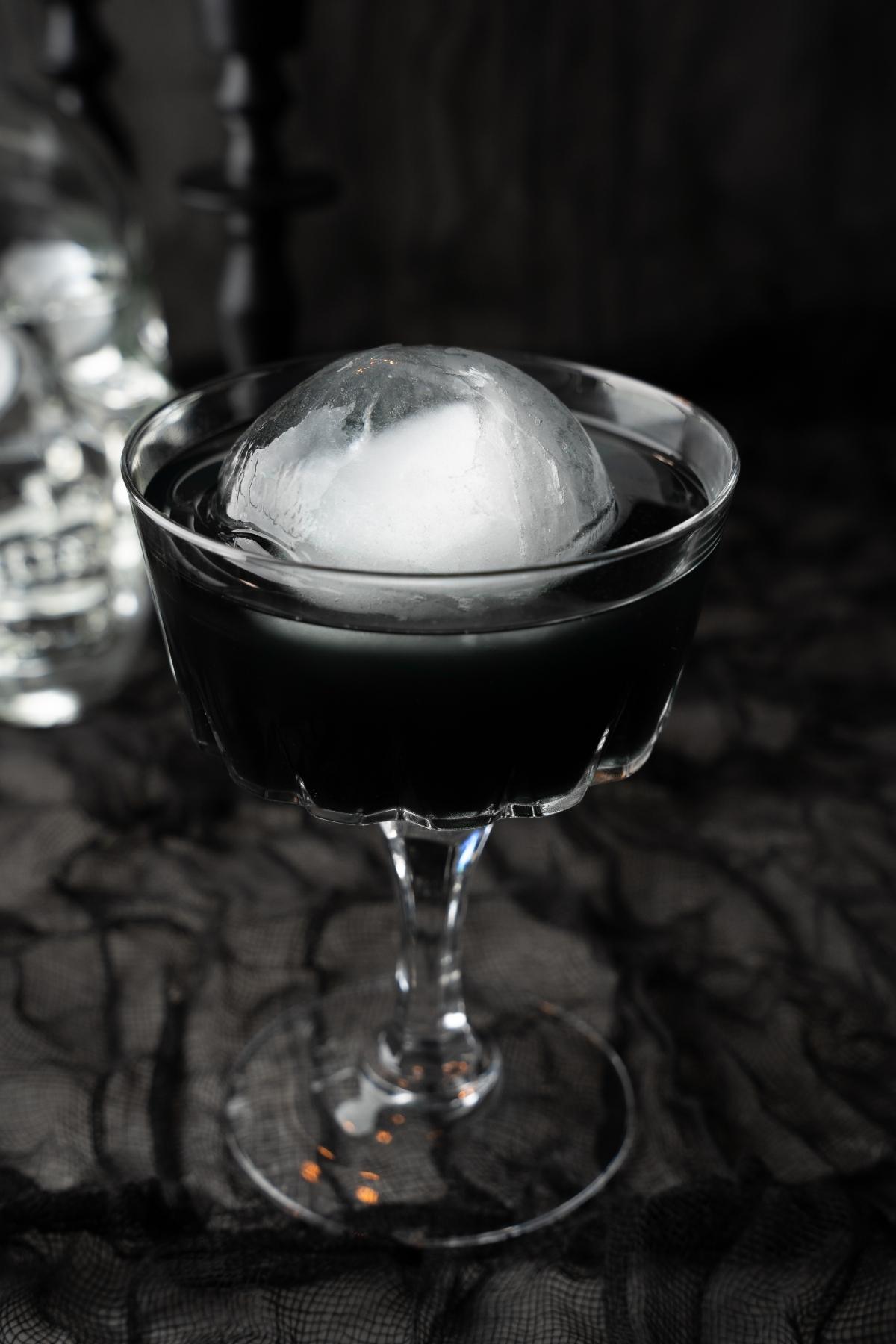 A Black Halloween Cosmopolitan Cocktail in a glass with a large round ice sphere rests on a black lacy surface, setting the perfect eerie mood.