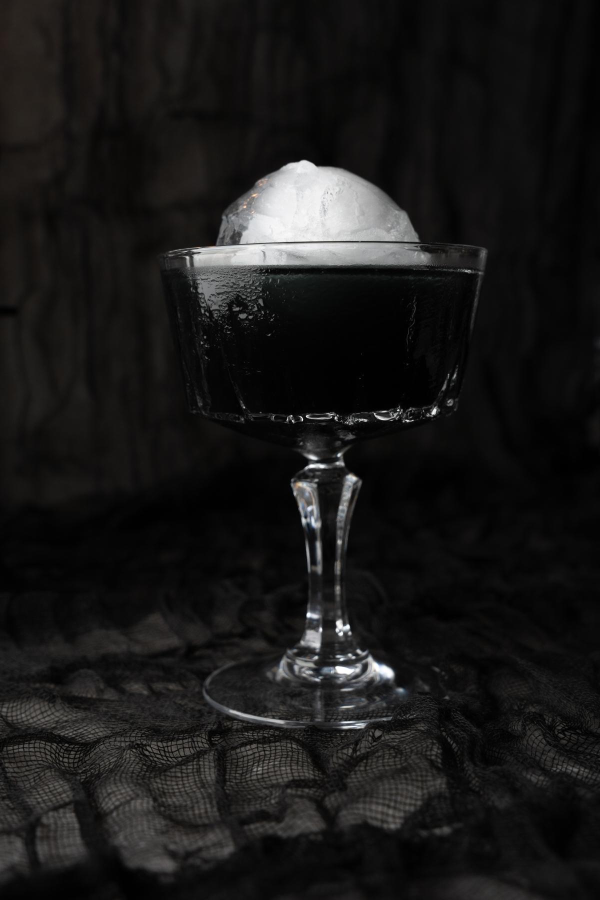 A black cocktail in a glass goblet with a large clear ice sphere, set against a black lace background—perfect as a Halloween themed drink.