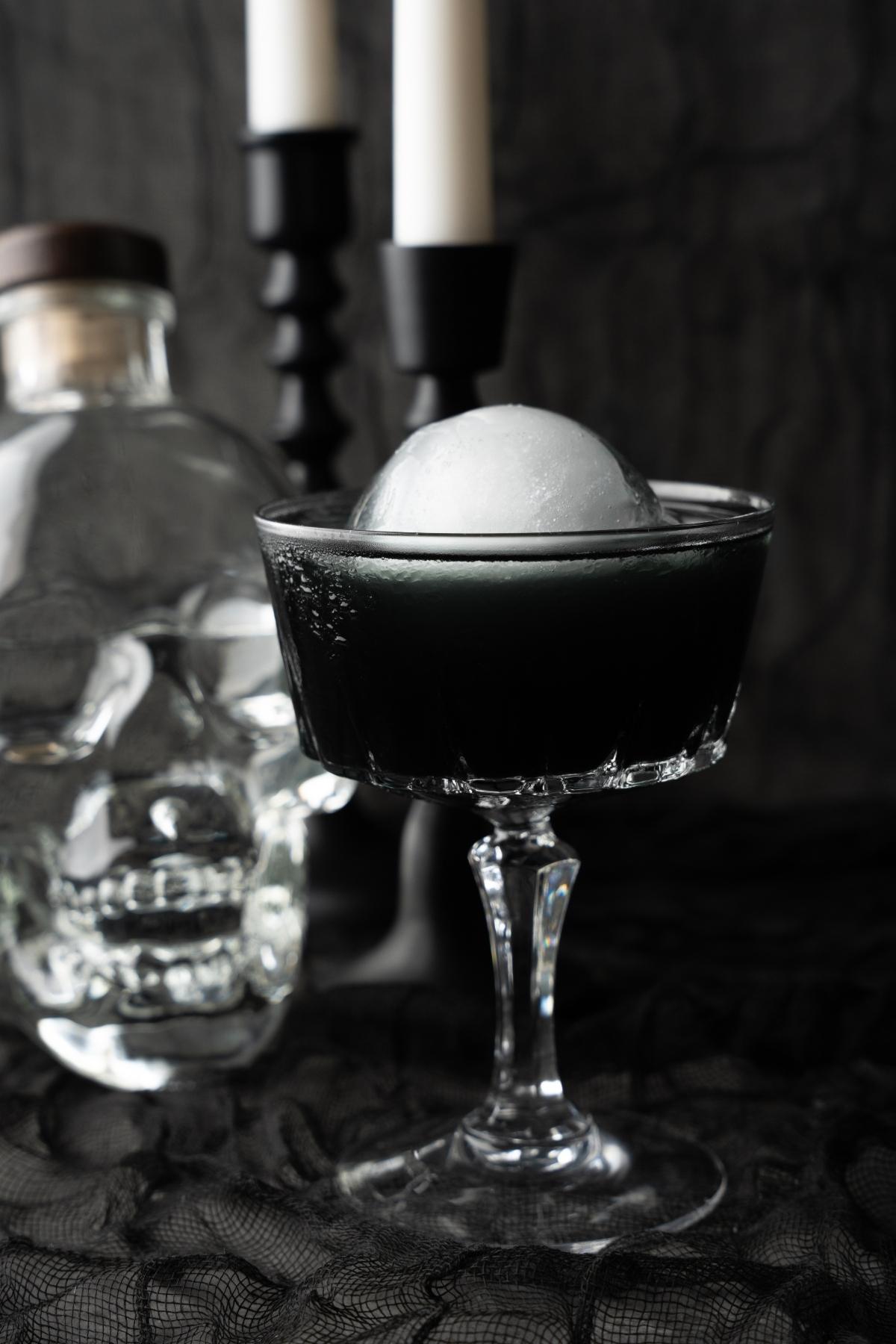 A mysterious black cocktail with a round ice ball sits in a glass beside a skull-shaped bottle and flickering candles, creating the perfect Halloween themed drink scene.