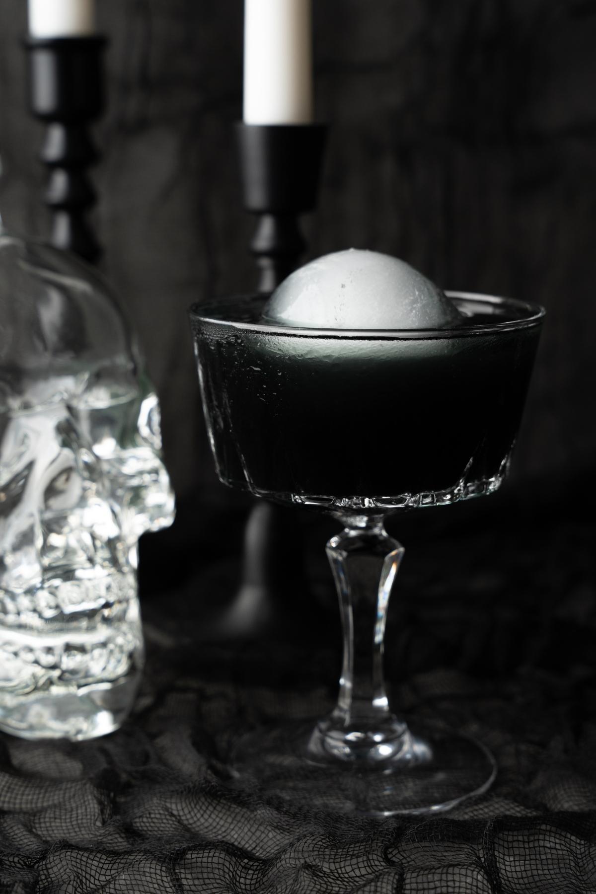 A striking black cocktail with a white ice sphere sits in a glass beside a glass skull and black candleholders, making it the perfect Halloween vodka drink.
