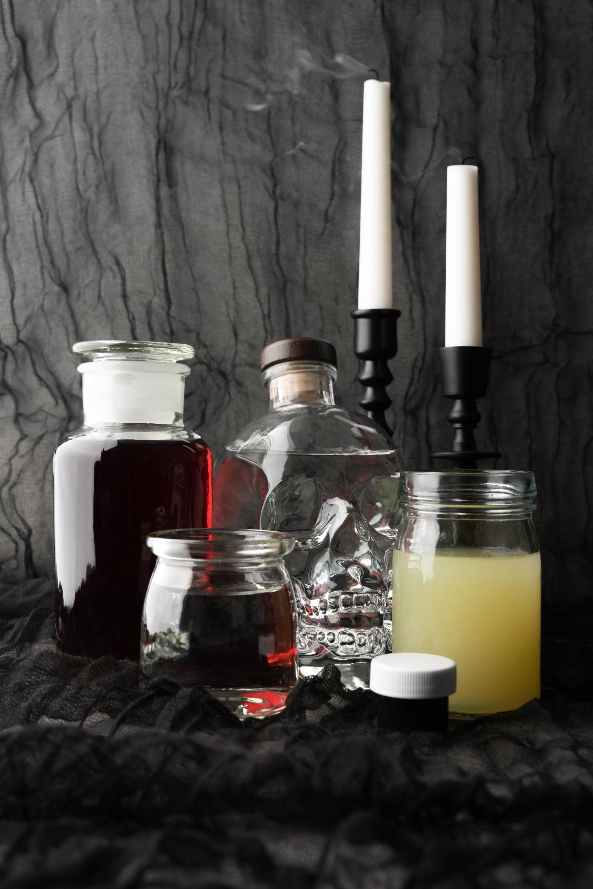 Assorted potion bottles and jars with candles on a dark lacy cloth evoke the magic of a Halloween themed drink, set against a textured gray background.