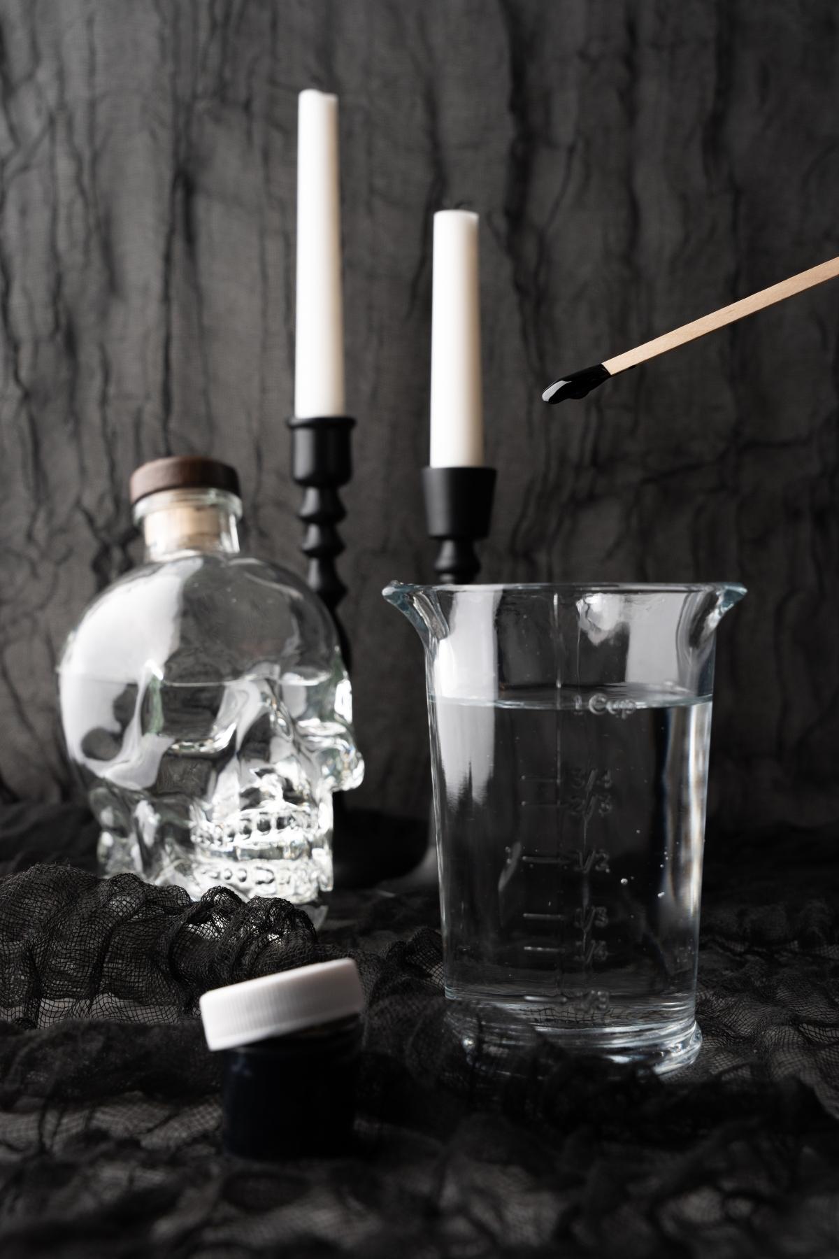 A clear beaker of water, a black cocktail on a stick, a skull-shaped bottle, candles, and a dark fabric background set the stage for this mysterious Black Halloween Cosmopolitan Cocktail.