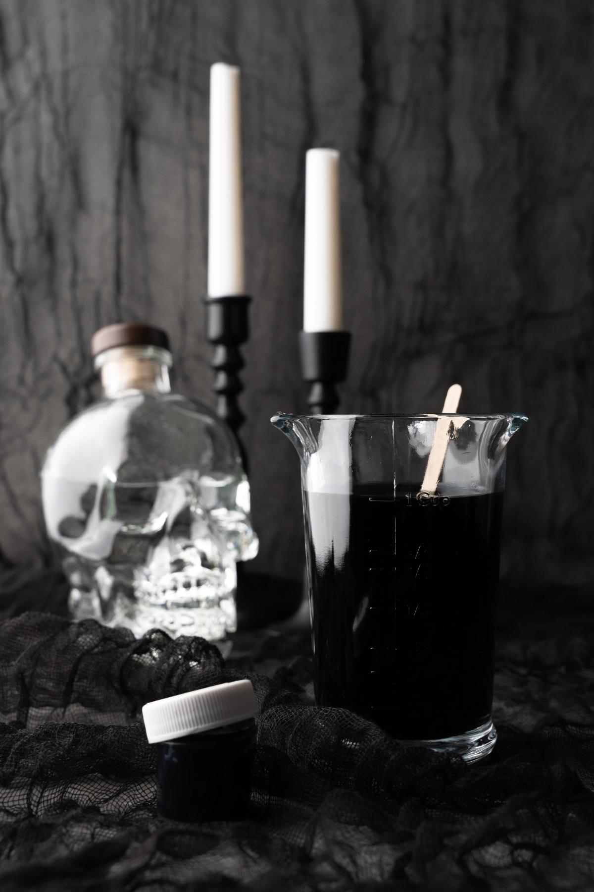 A glass of black cocktail, a skull-shaped bottle, and two white candles create a mysterious scene on a dark, textured background—perfect for a spooky Halloween vodka drink.