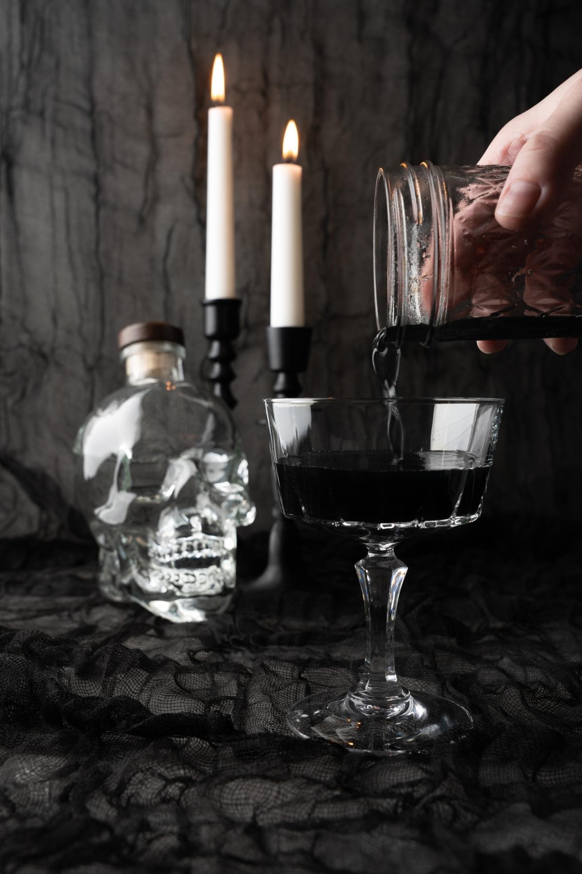 A hand pours a black cocktail with an ice ball into a glass, with a skull bottle and lit candles in the background.