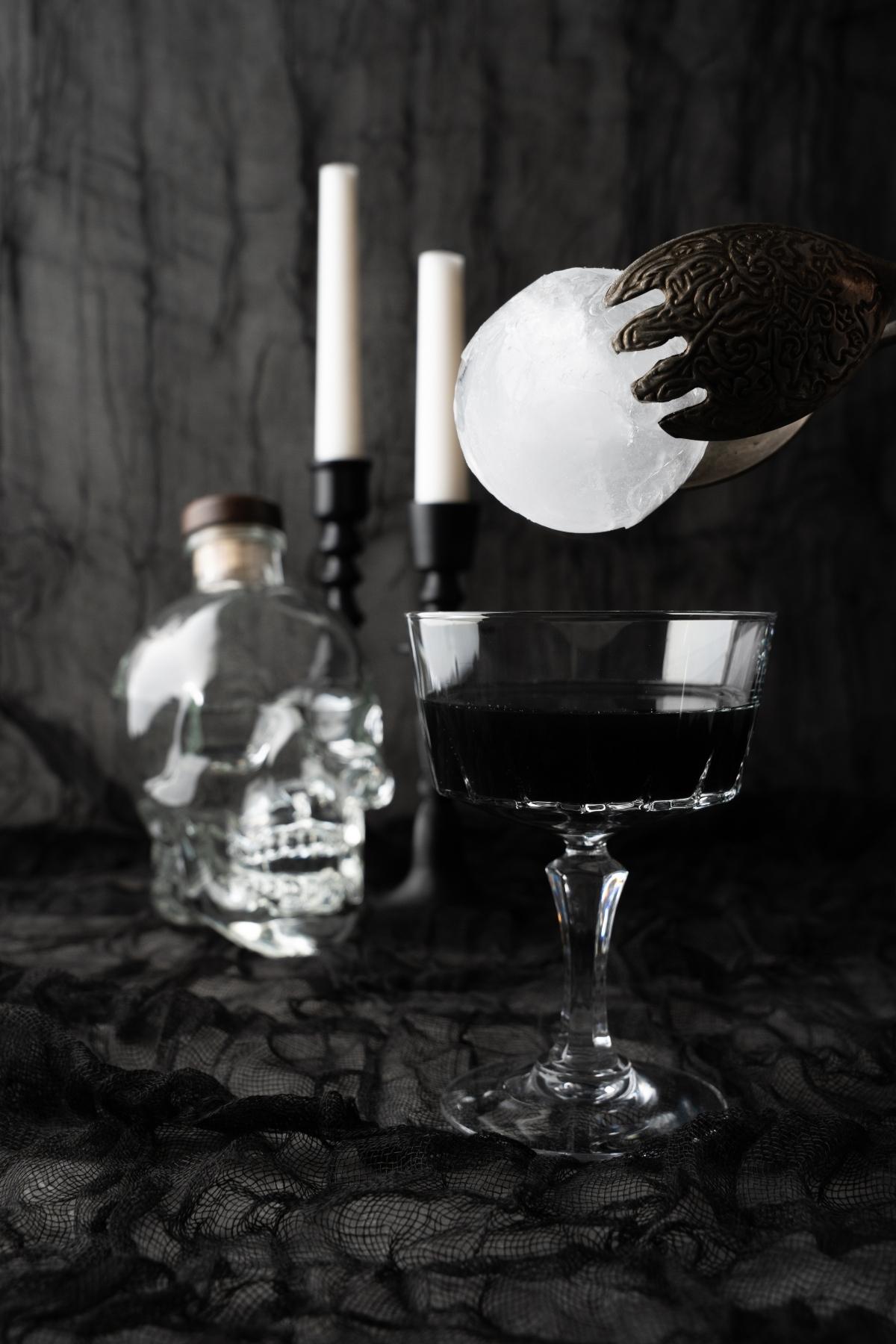A claw holds a round ice sphere above a Black Halloween Cosmopolitan Cocktail, with a skull bottle and candles in the background, setting the perfect scene for a Halloween themed drink.