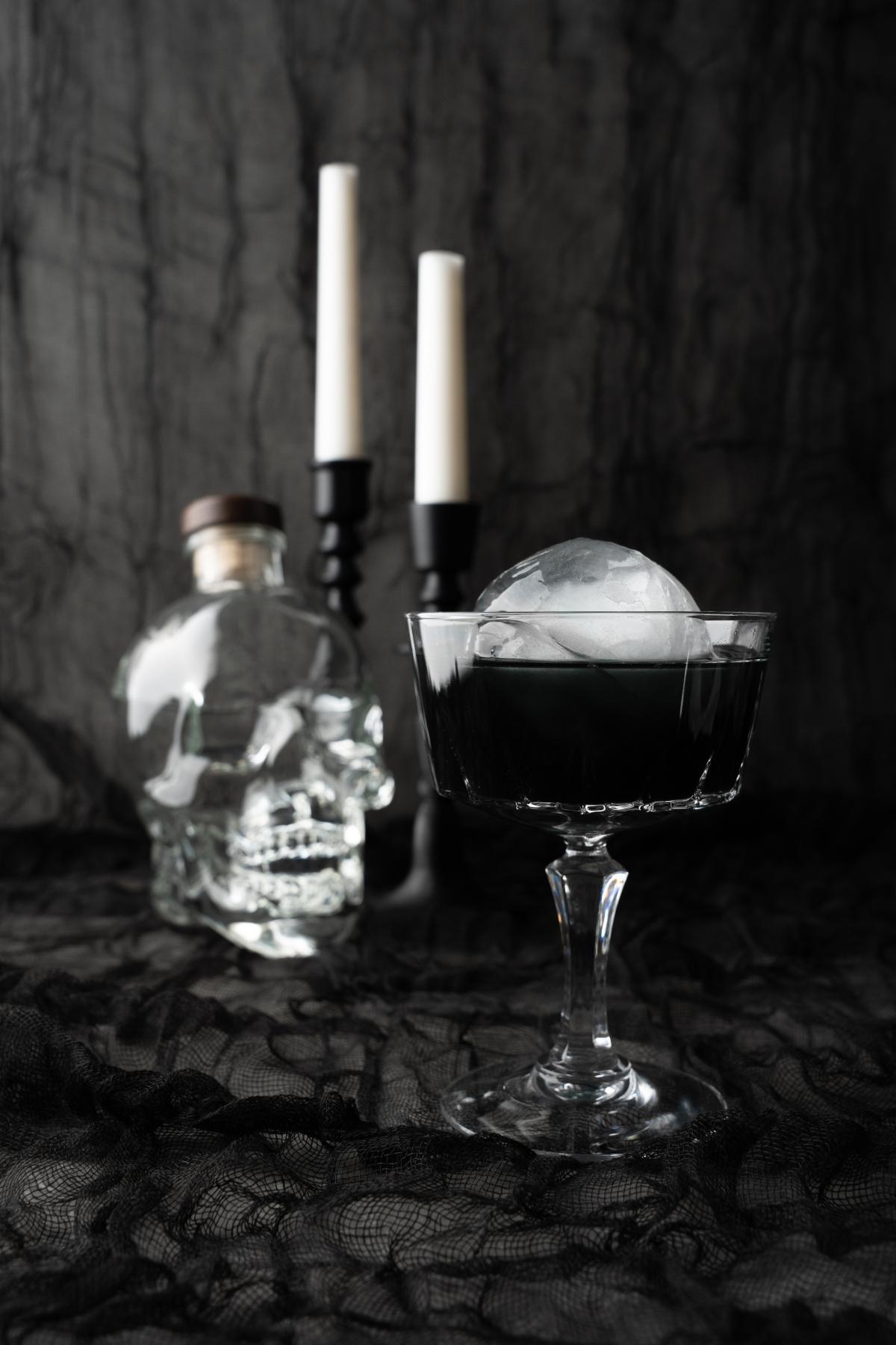 A dark Black Halloween Cosmopolitan Cocktail with a round ice cube, a skull-shaped bottle, and two candles rests on a black lace cloth.