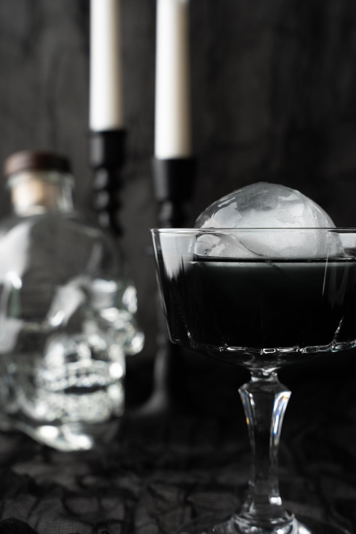 A dark Black Halloween Cosmopolitan Cocktail with a large ice sphere sits in a glass, while a skull-shaped bottle and flickering candles create the perfect spooky backdrop for this Halloween vodka drink.