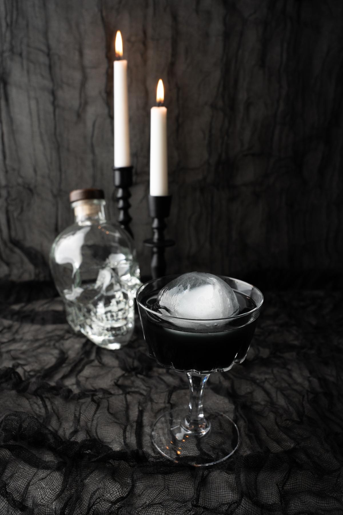 A black Halloween Cosmopolitan cocktail with a round ice ball sits beside a skull-shaped bottle and two lit candles on a black cloth—an eerie, Halloween themed drink for spooky nights.