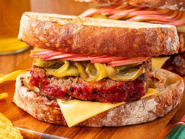 A hearty meatloaf sandwich with cheese, pickles, and crisp apple slices on rustic bread, served alongside crunchy potato chips.