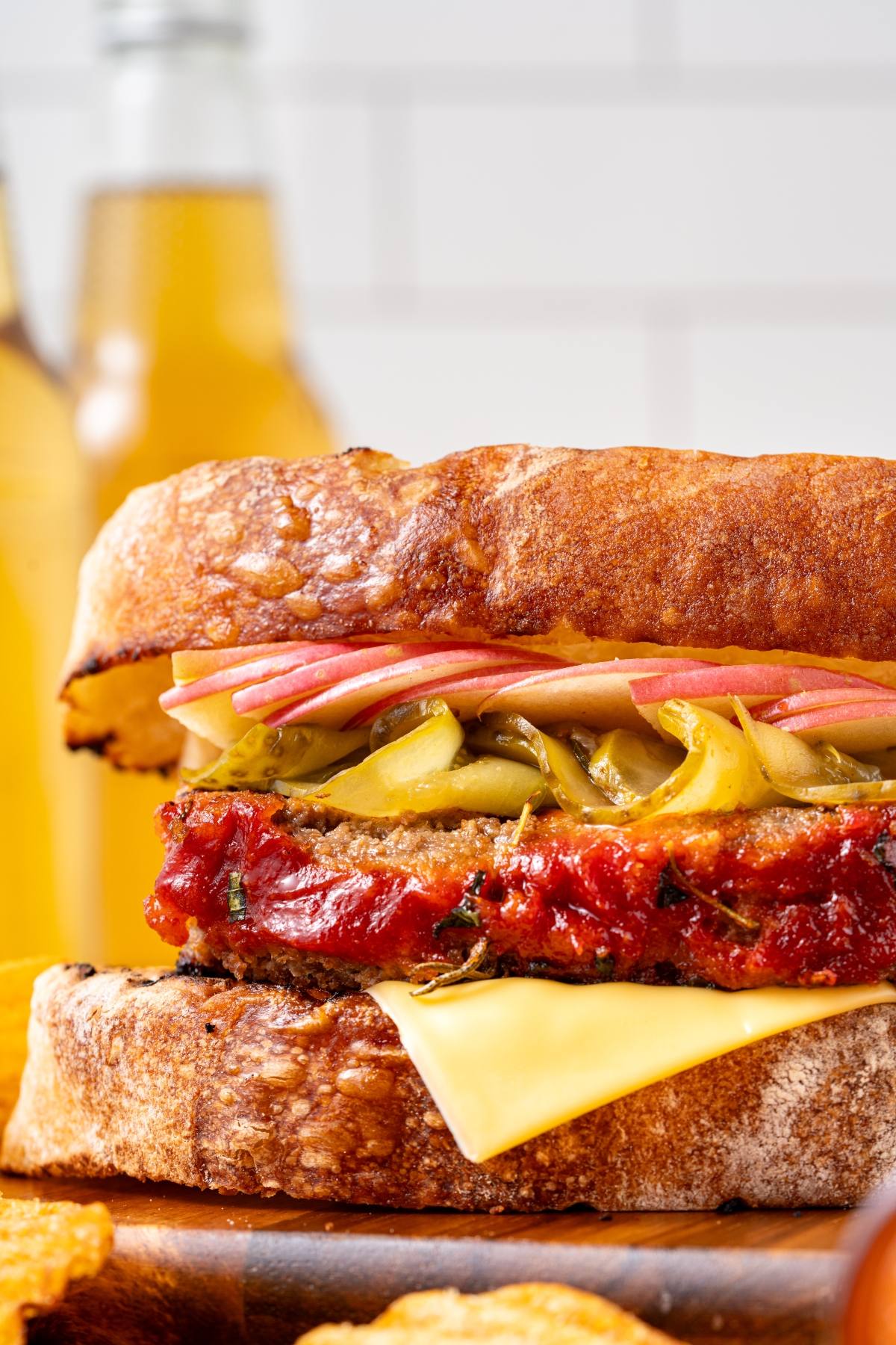 Close-up of a sandwich with meatloaf, cheese, pickles, ham, and mustard on rustic bread; drinks in background.