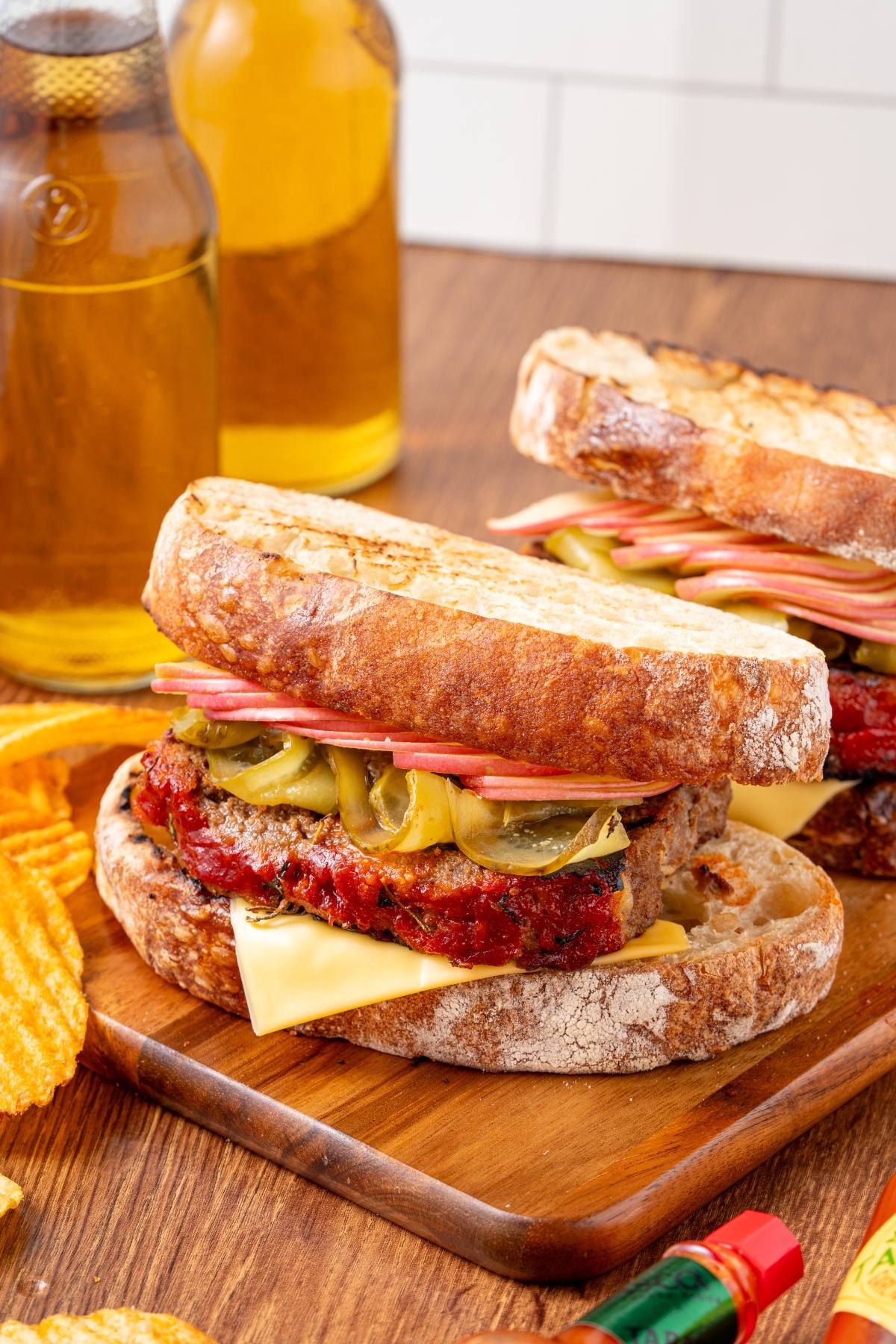 Two meatloaf sandwiches with cheese, pickles, and tomato on rustic bread, served with chips and drinks.