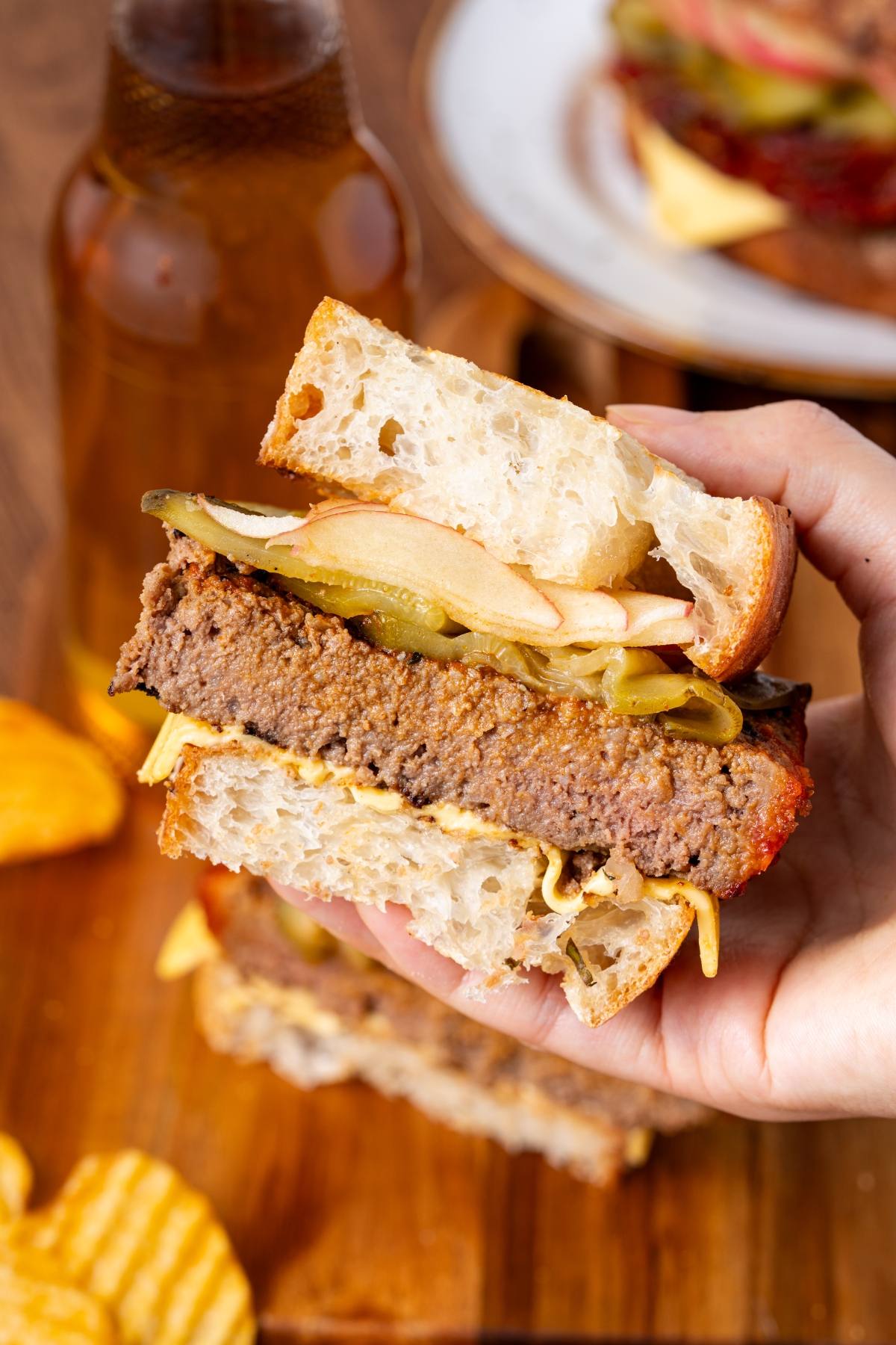 A hand holds a sandwich with thick bread, beef patty, pickles, and cheese; chips and a drink nearby.