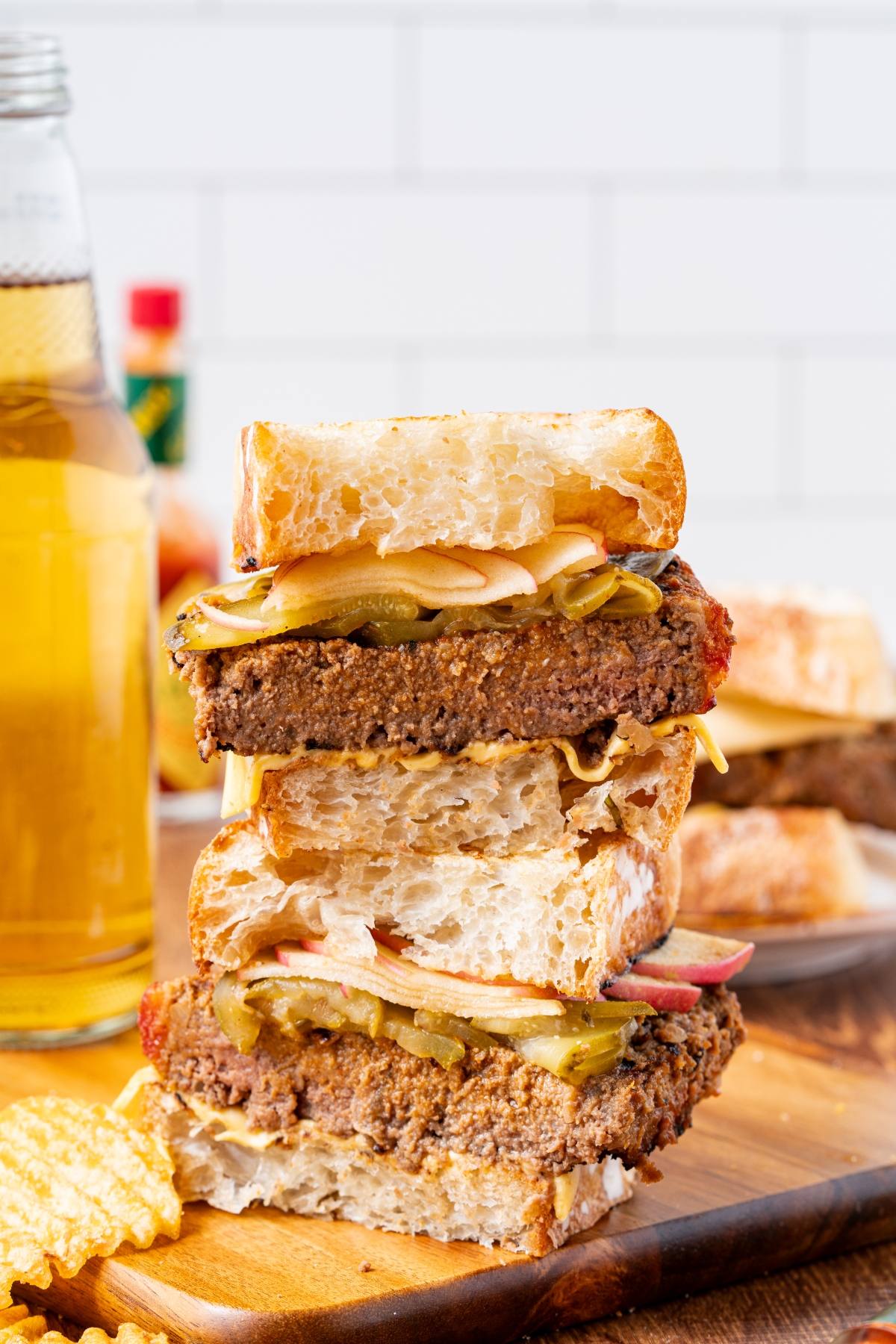 A stacked sandwich with bread, cheese, pickles, apples, and a burger patty, next to chips and a drink.