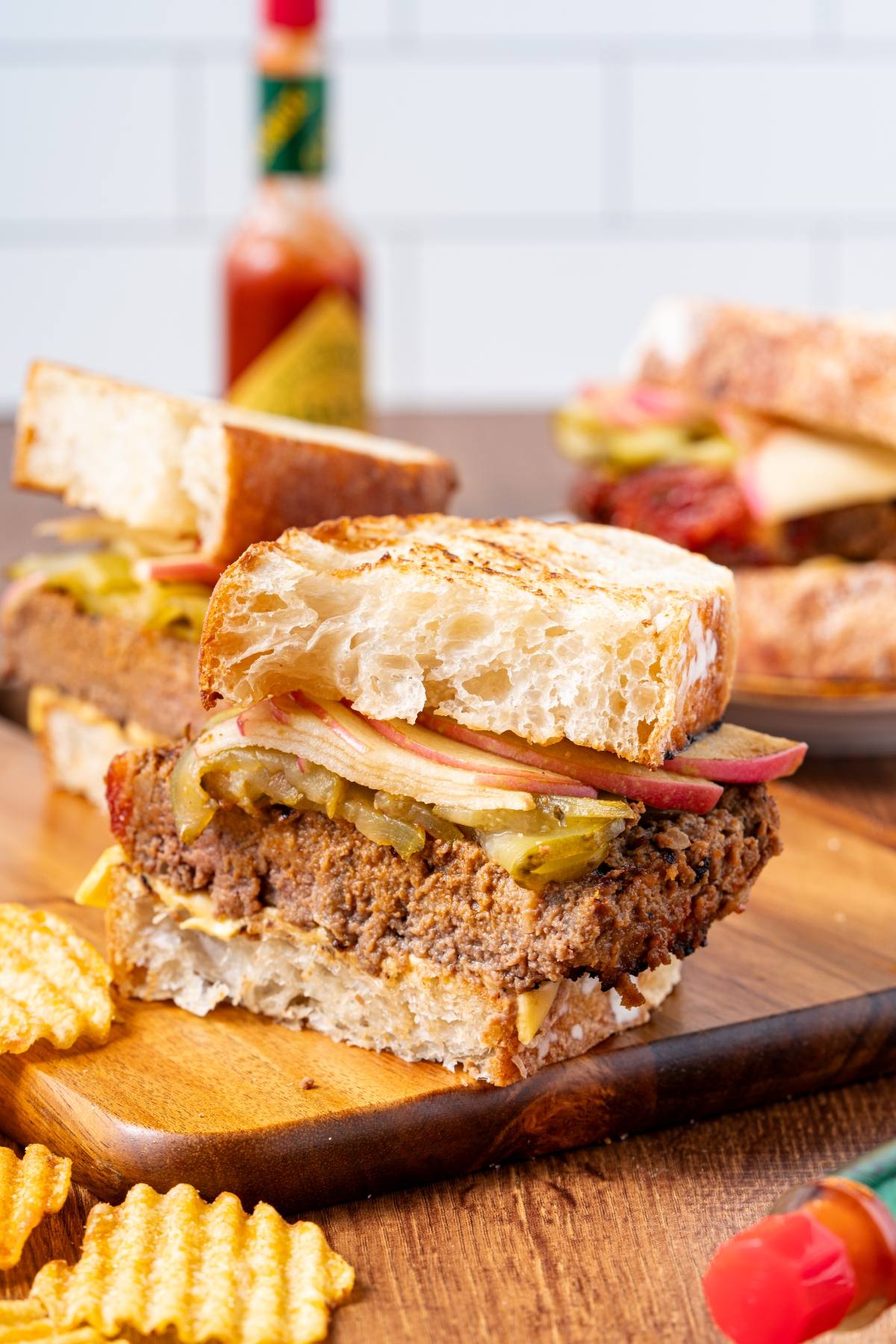 A meatloaf sandwich with pickles on rustic bread, served on a wooden board with crinkle-cut potato chips.
