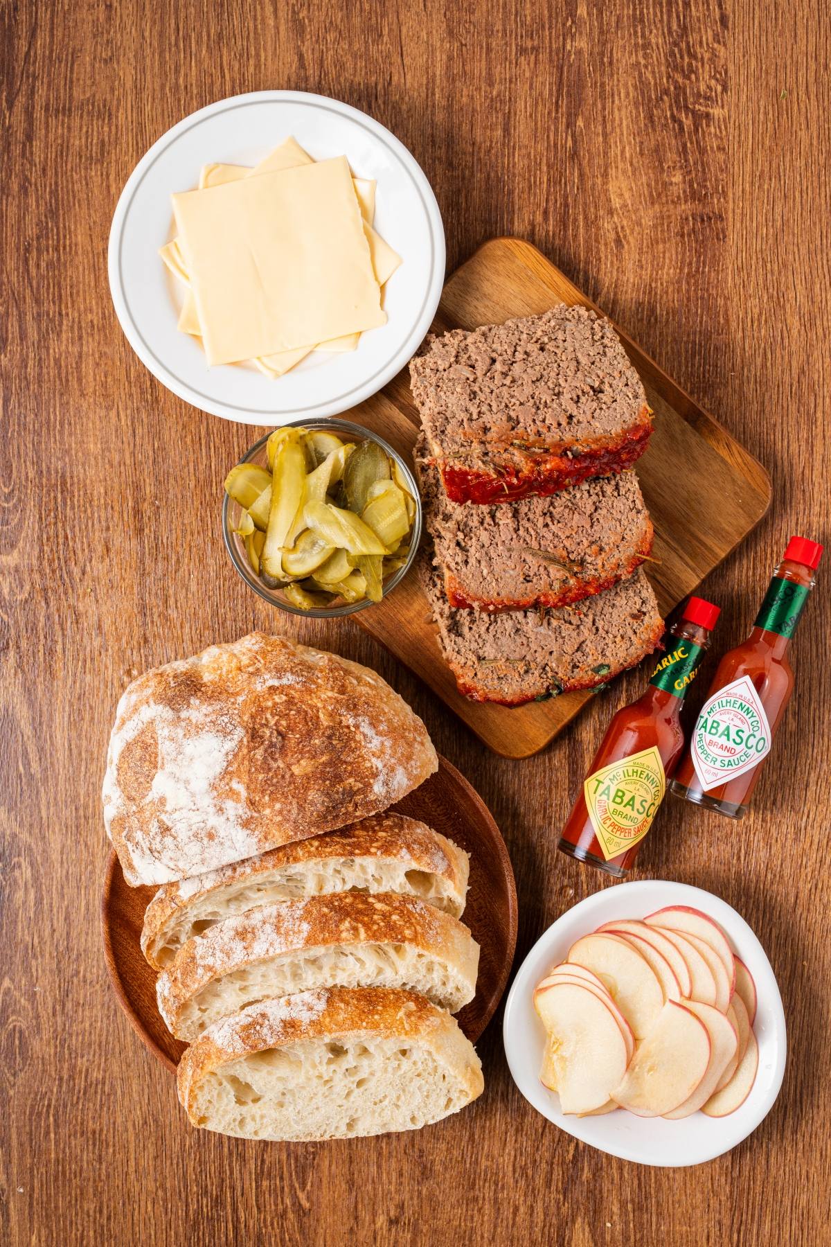 Sliced bread, cheese, meatloaf, pickles, apple slices, and Tabasco bottles on a wooden table.