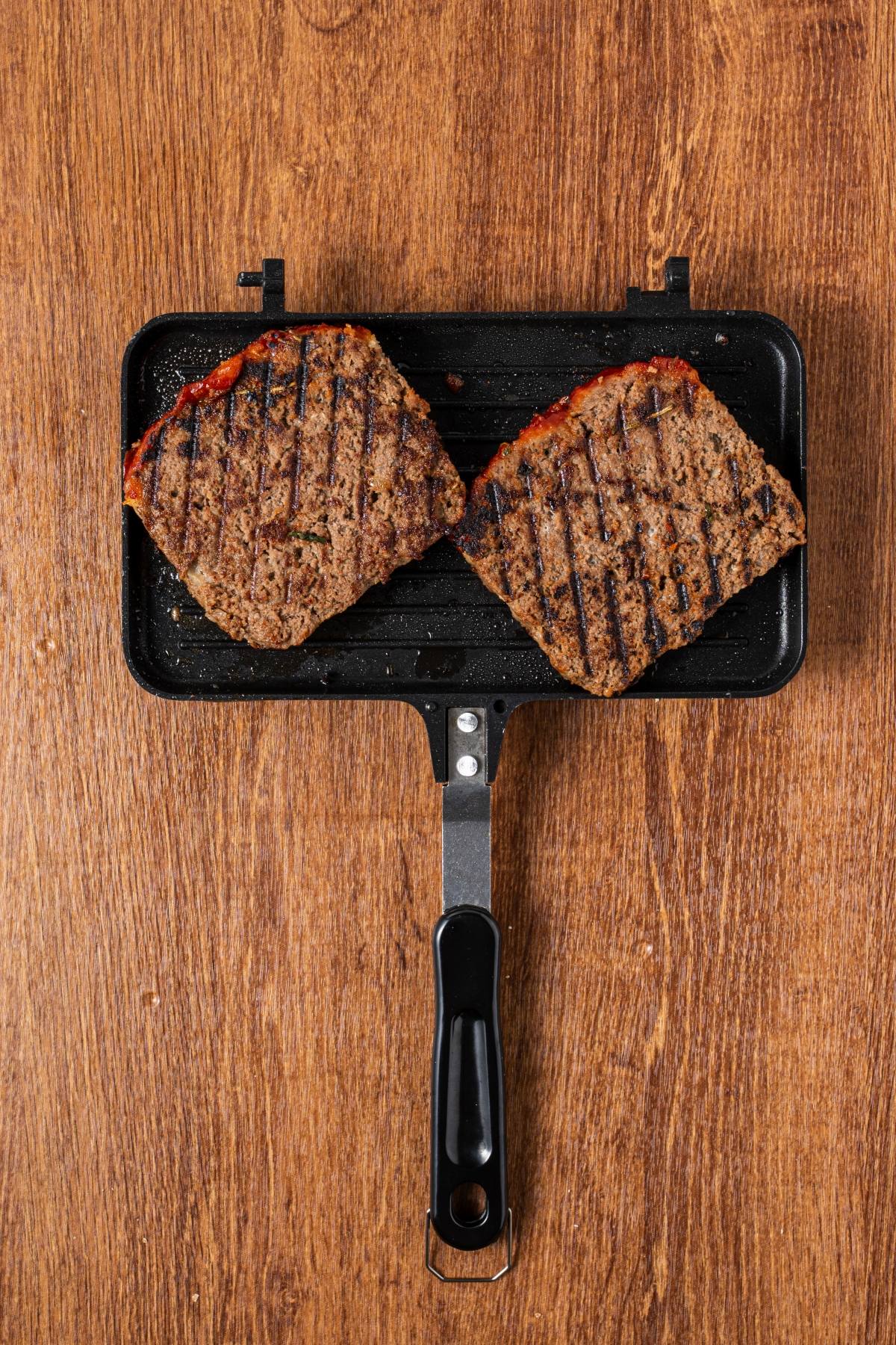 Two grilled meatloaf slices with char marks sit in a rectangular grill pan on a wooden surface.