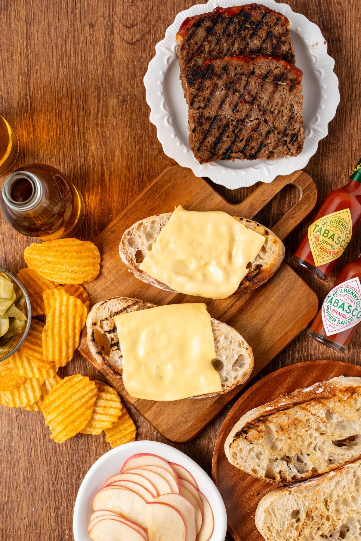 Sliced bread with cheese, grilled meatloaf, crinkle chips, apple slices, pickles, and Tabasco sauce on a table.