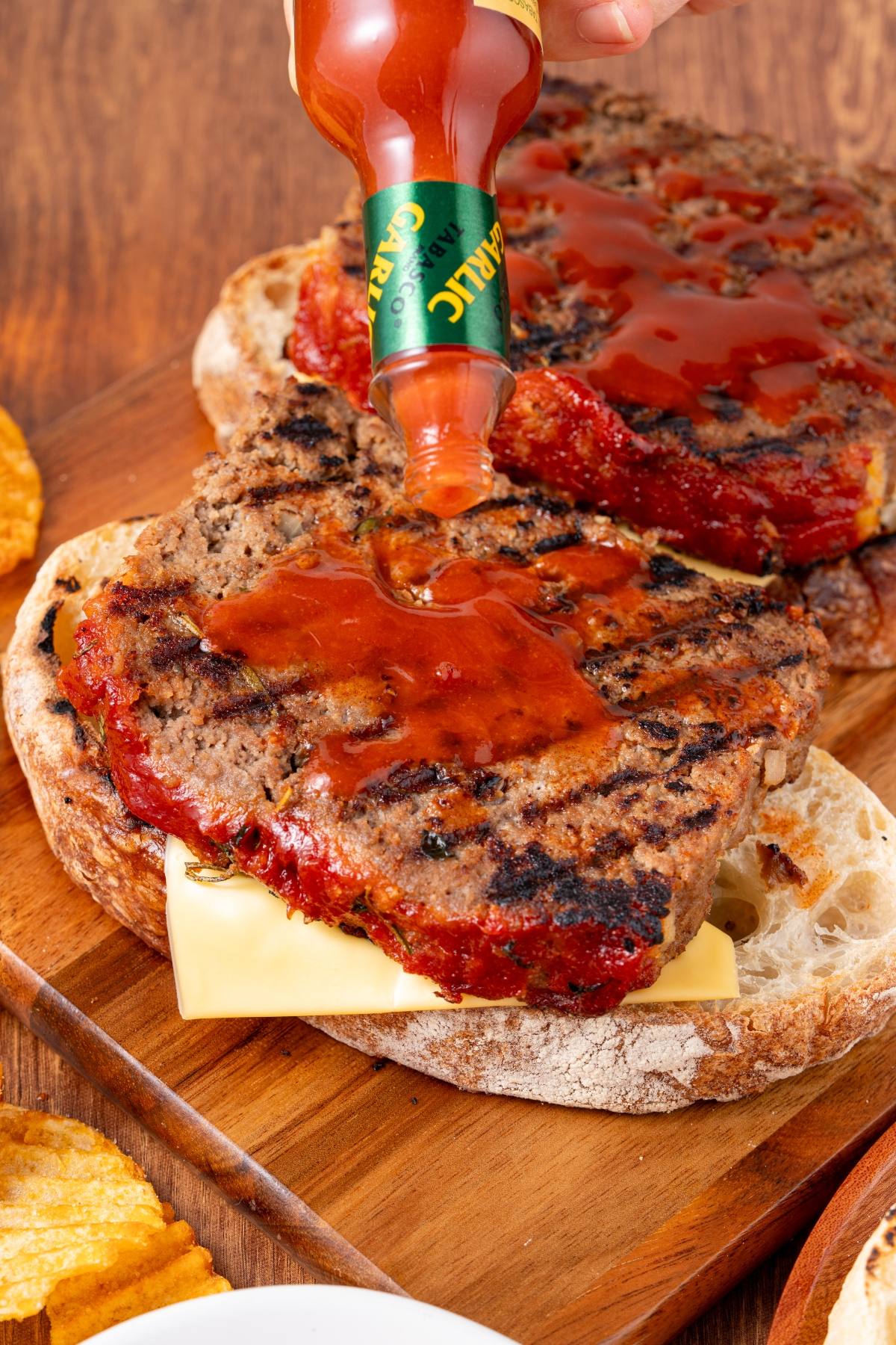A grilled meatloaf sandwich on bread with cheese, topped with hot sauce being poured from a bottle.