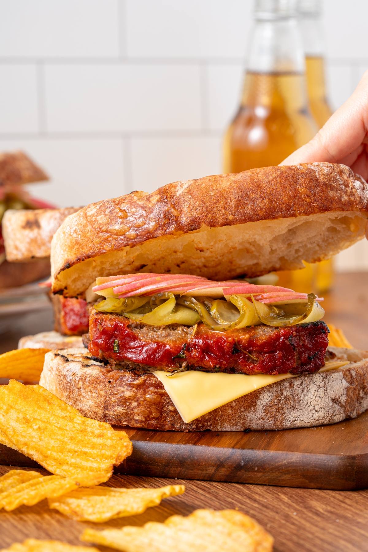 A hand lifts the top slice of bread on a meatloaf sandwich with cheese, pickles, onions, and chips on the side.