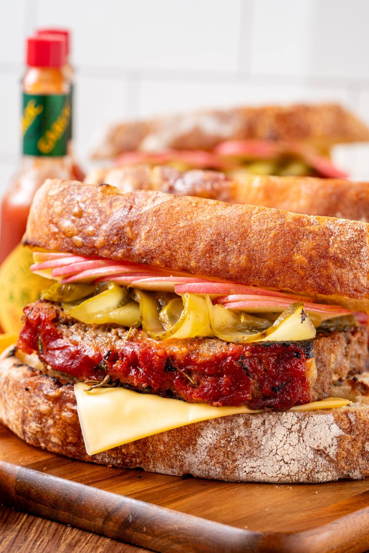 A close-up of a stacked sandwich with cheese, meat, pickles, and sauce on rustic bread, with hot sauce in the background.