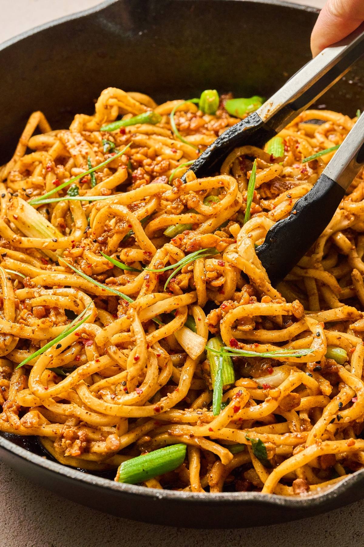 Tongs mixing spicy noodles with ground meat and green onions in a skillet.