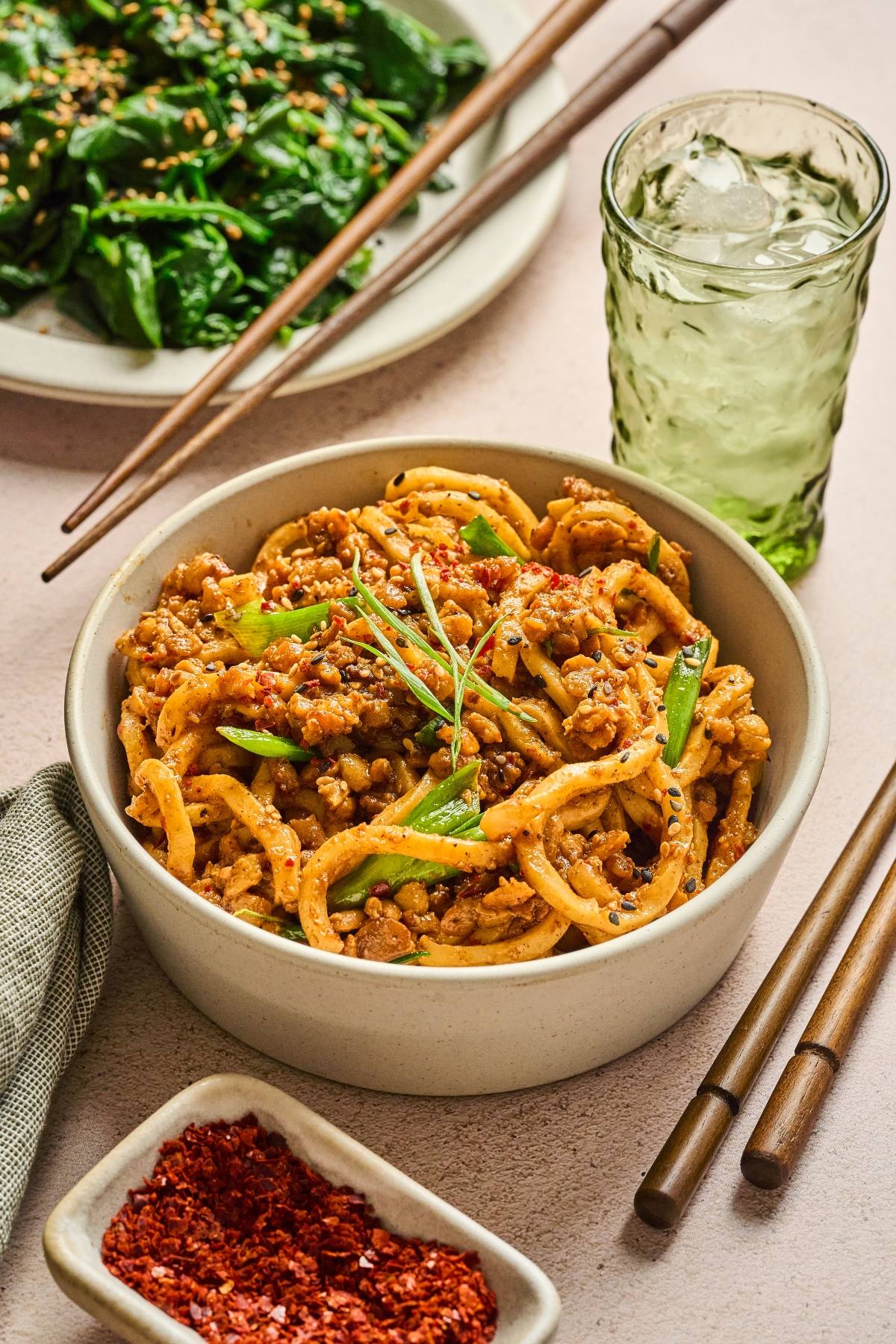 A bowl of spicy noodles with ground meat and green onions, chopsticks, and a glass of iced drink nearby.