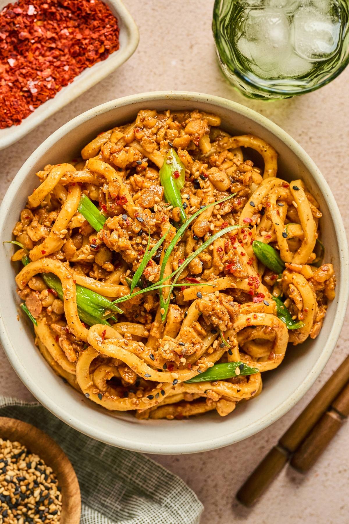 A bowl of spicy noodles with ground meat, green onions, and sesame seeds, with seasonings on the side.