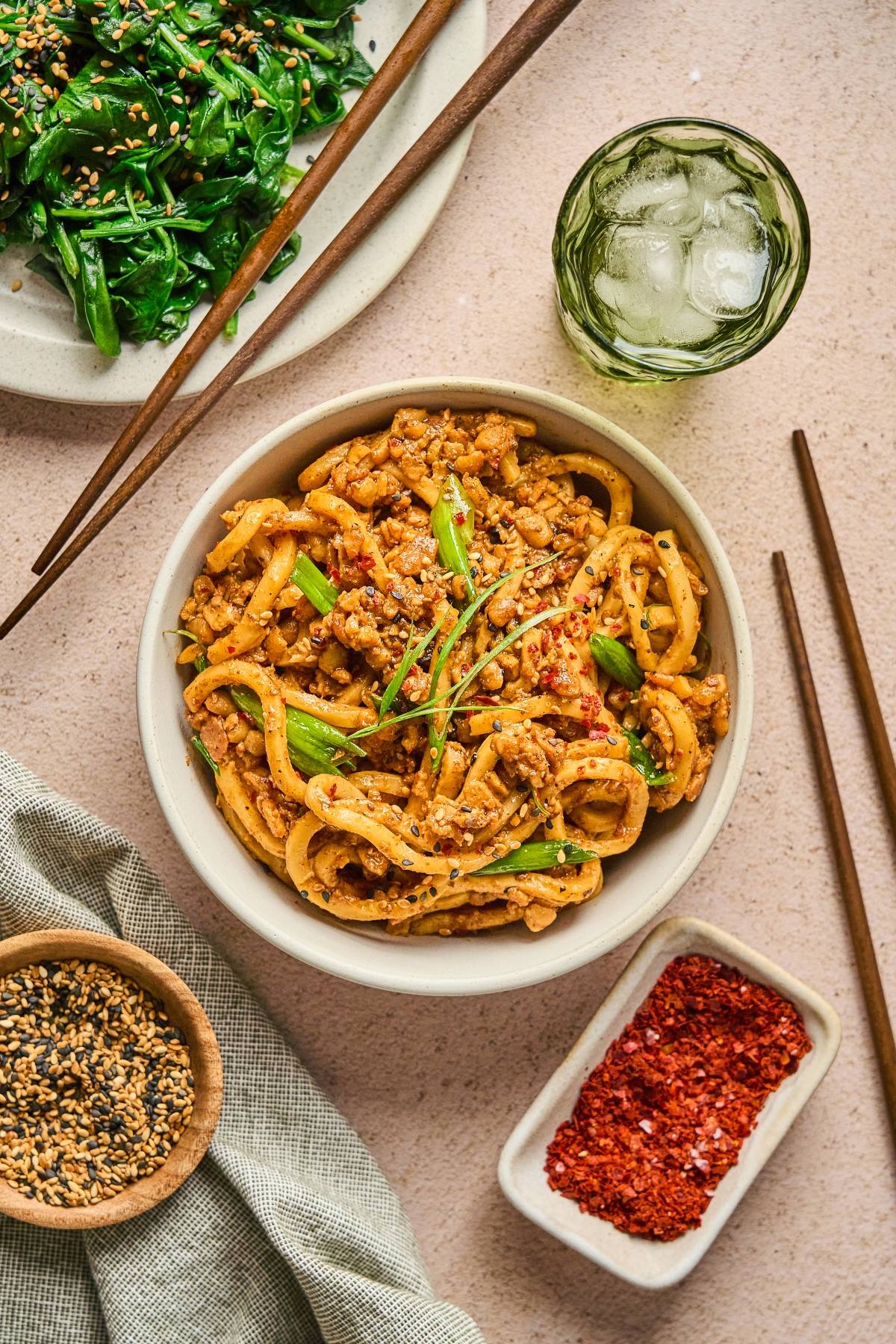 A bowl of spicy noodles with ground meat and green onions, surrounded by side dishes, chopsticks, and a drink.