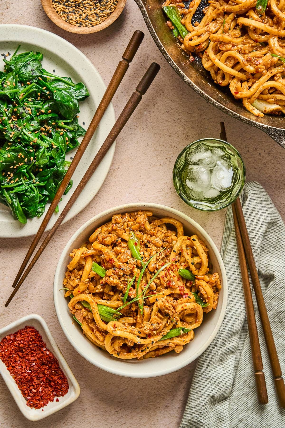 Bowl of spicy noodles with ground meat, fresh greens, chopsticks, ice water, and seasonings on a table.