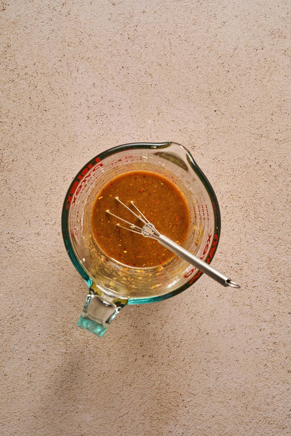 Glass measuring cup with brown liquid and a small whisk on a light textured surface, viewed from above.