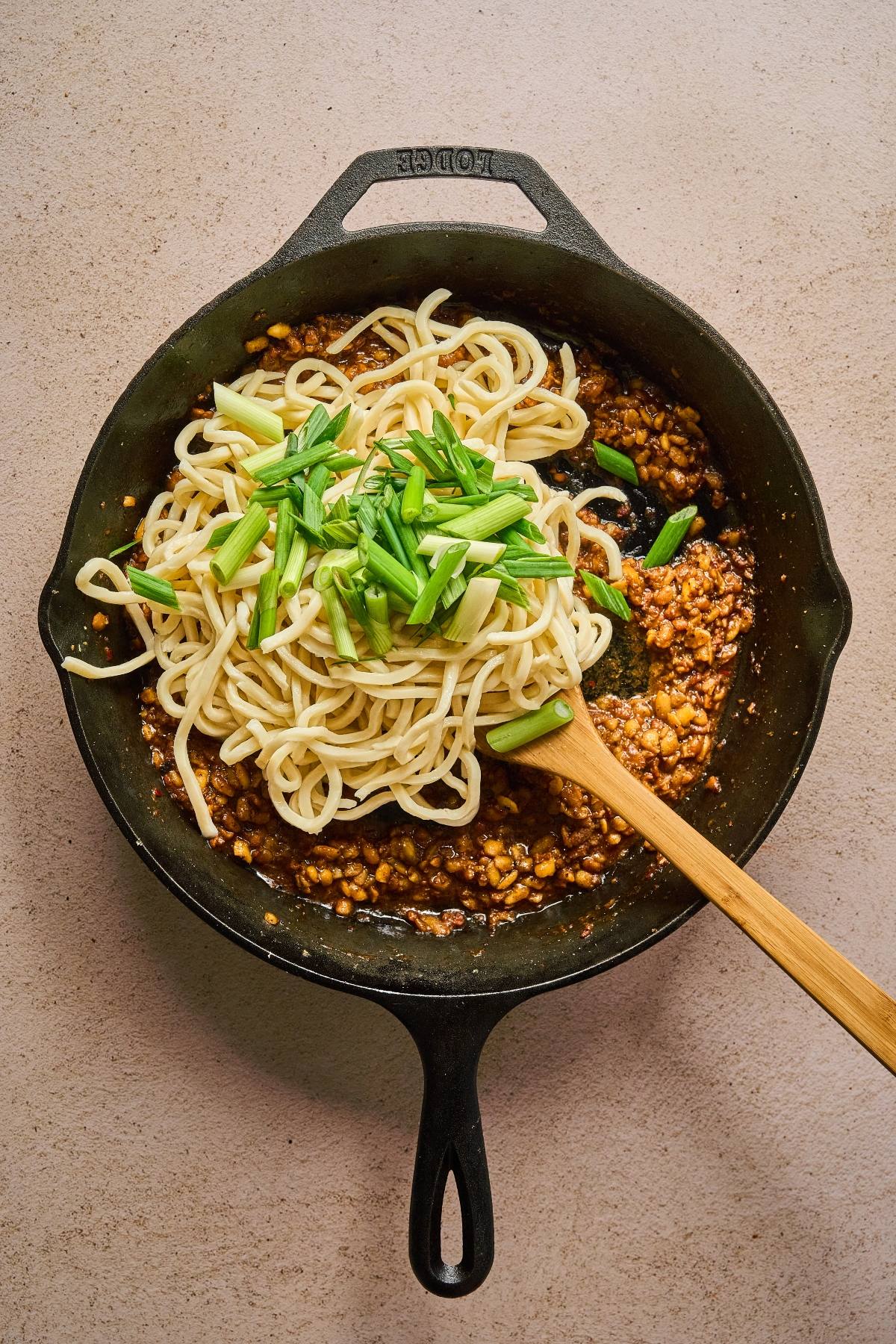 A cast iron skillet with noodles, chopped green onions, and a savory sauce being mixed with a wooden spoon.
