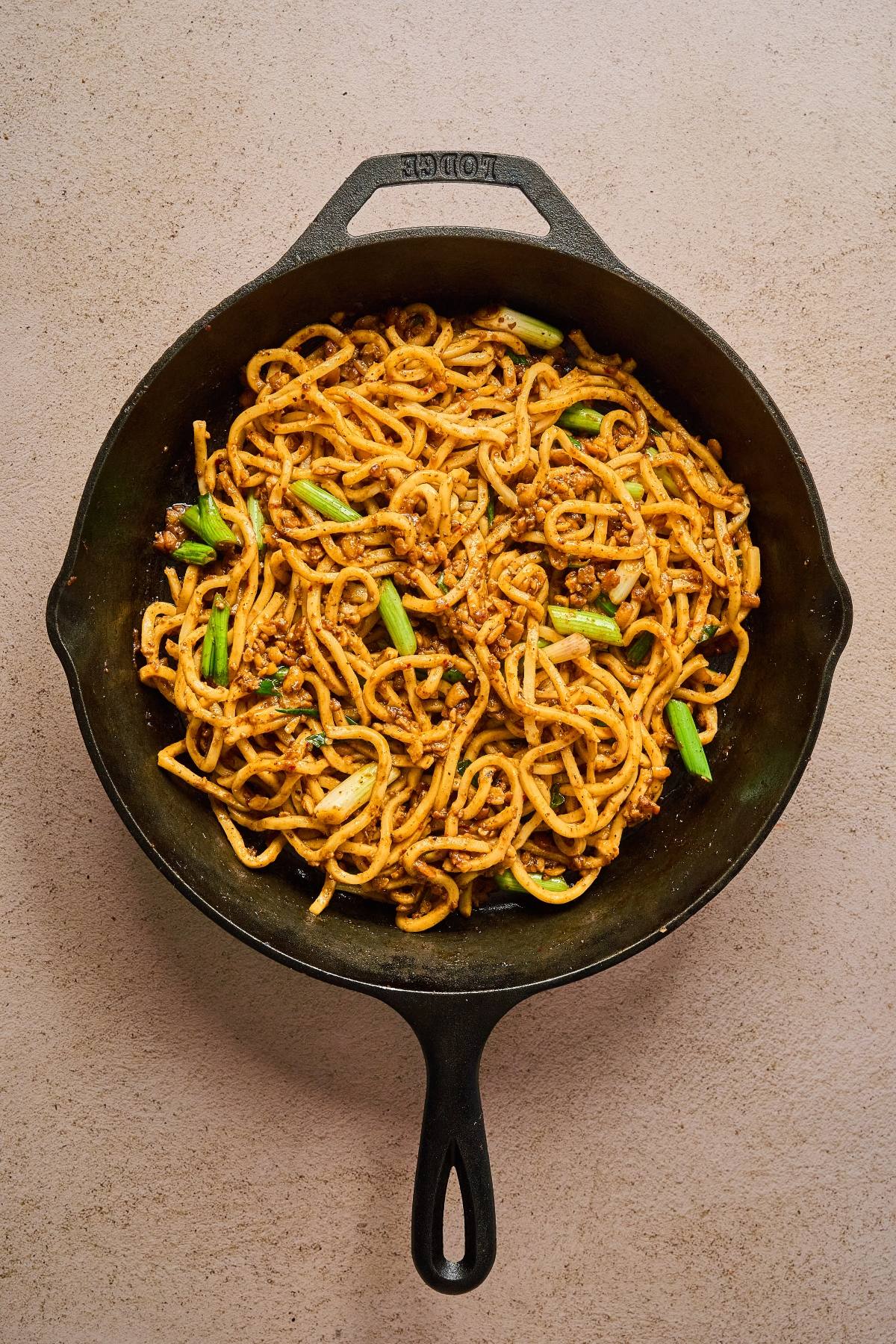 A cast iron skillet filled with stir-fried noodles, ground meat, and chopped green onions.
