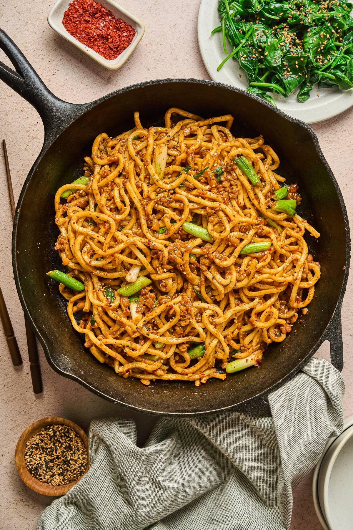 A cast iron skillet of stir-fried noodles with vegetables, surrounded by sides and chopsticks on a table.